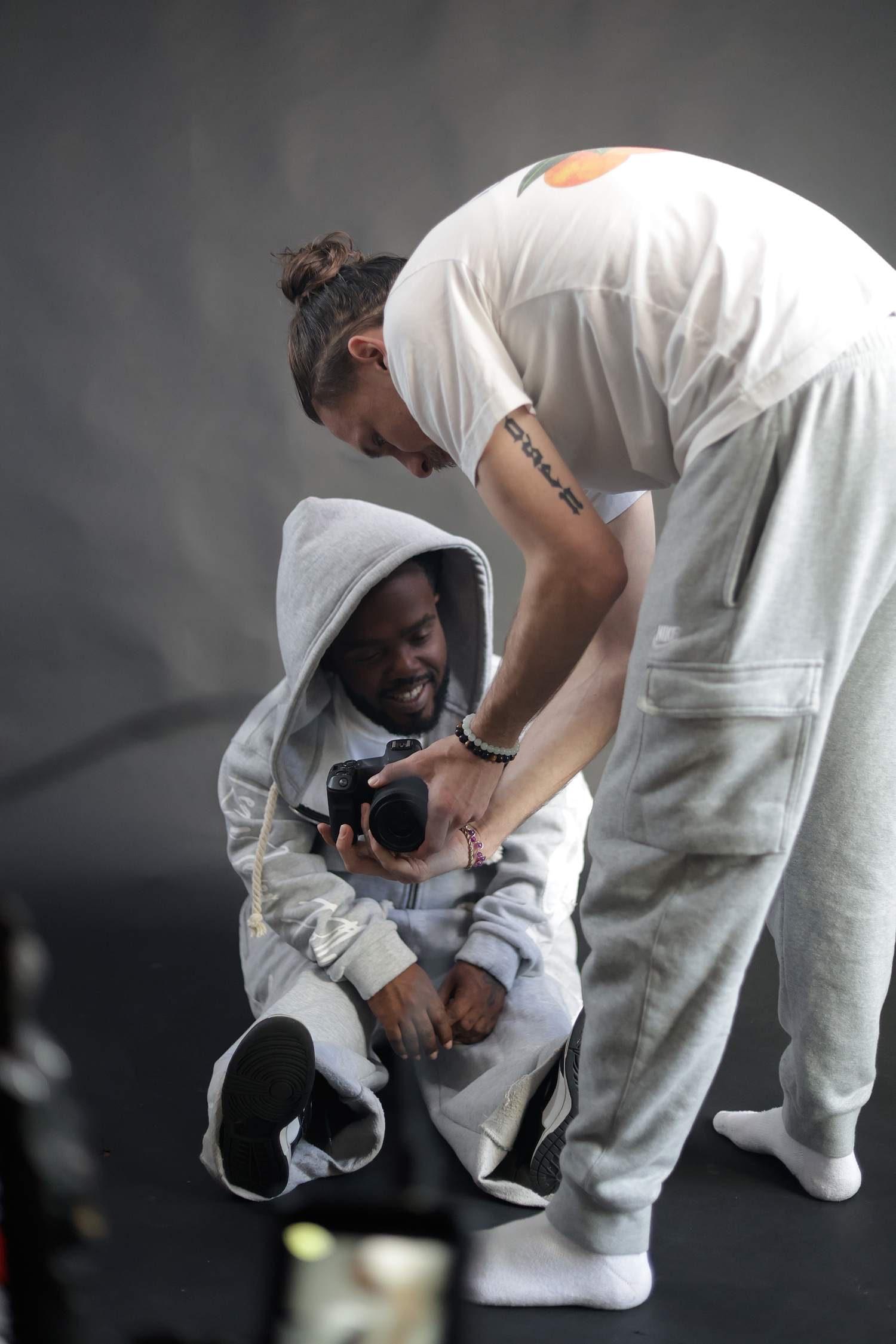Photographer shows camera preview to seated subject wearing grey hoodie in dark studio.