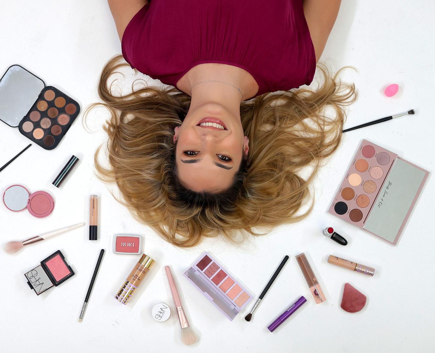 Smiling woman with long wavy hair lying on her back, surrounded by makeup palettes, brushes, lipsticks and blush on a white background.