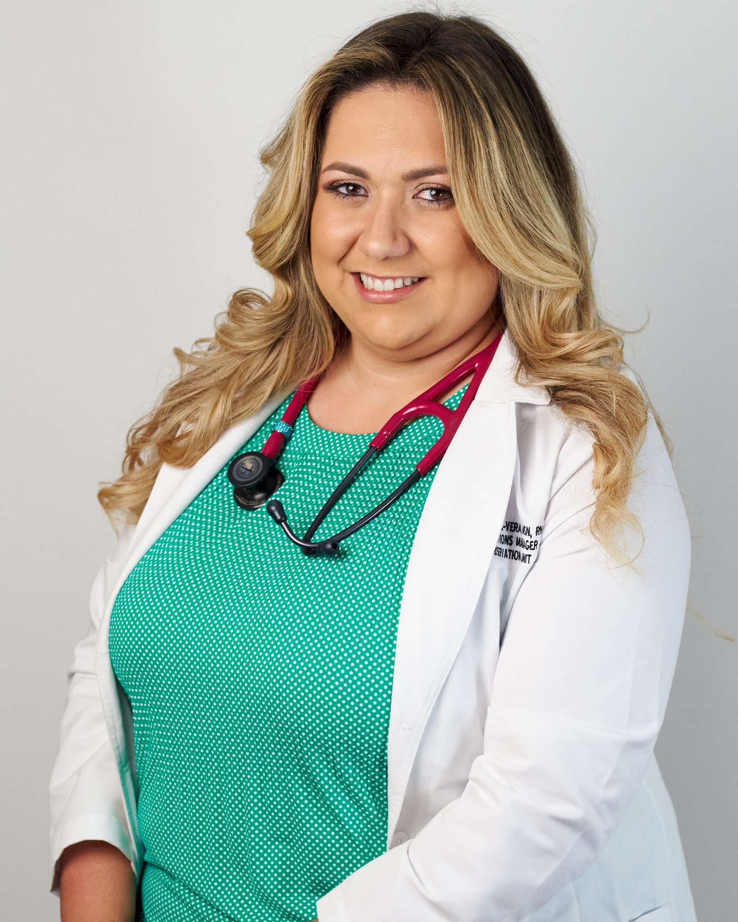 Smiling female medical professional in white coat and green top with red stethoscope in studio portrait.