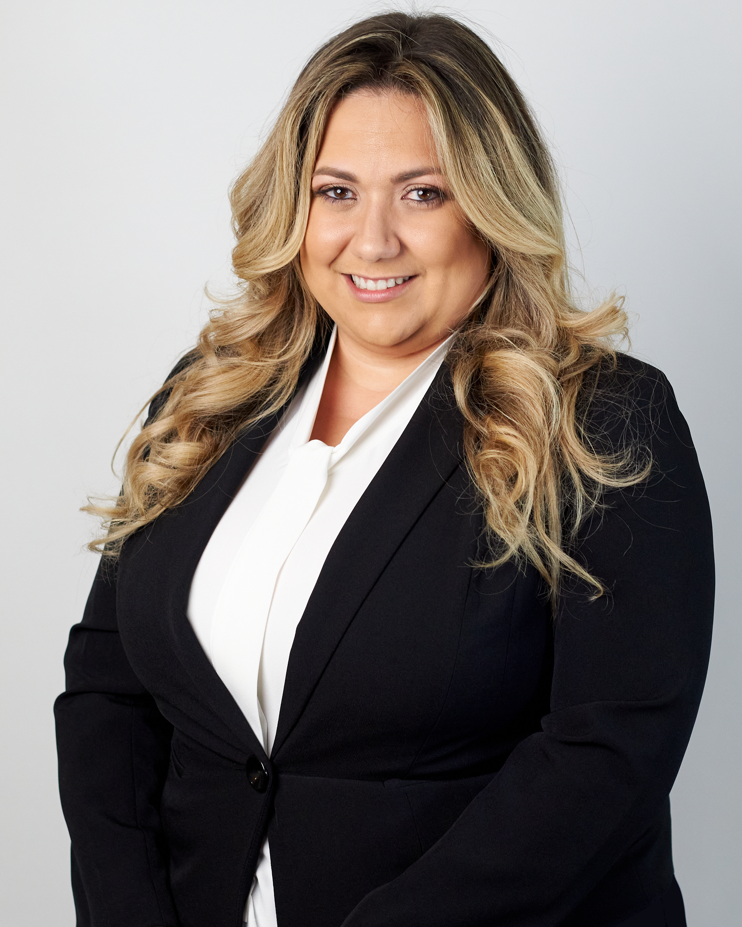 Professional woman with blonde wavy hair smiles in black blazer and white blouse for business portrait.
