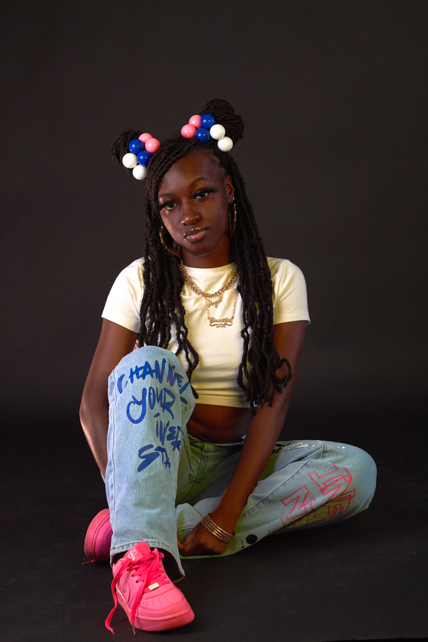 Young Black woman with braided hair and colorful pom-poms sitting cross-legged in yellow crop top and jeans.