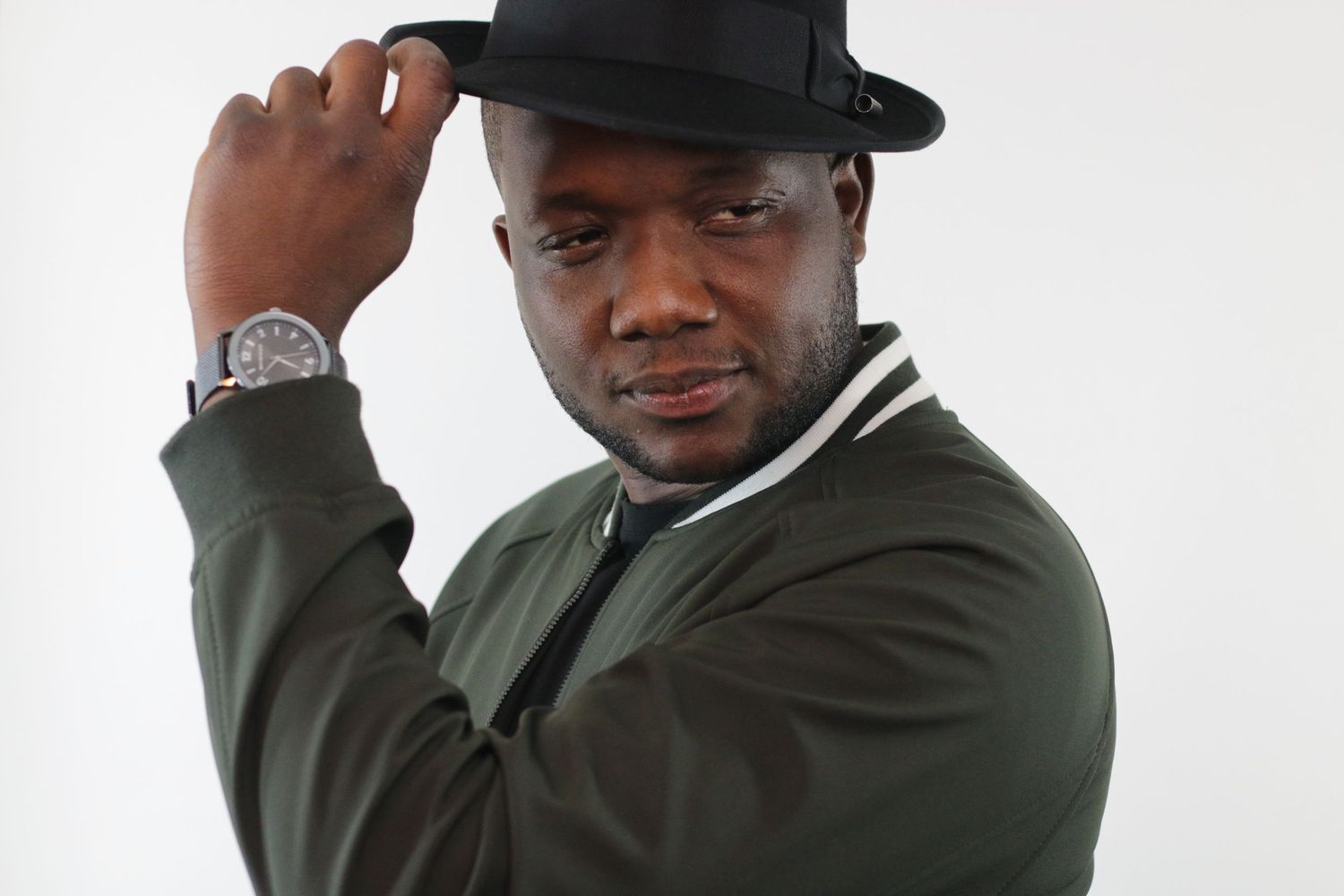 Confident man in olive jacket and black fedora hat posing against white background.