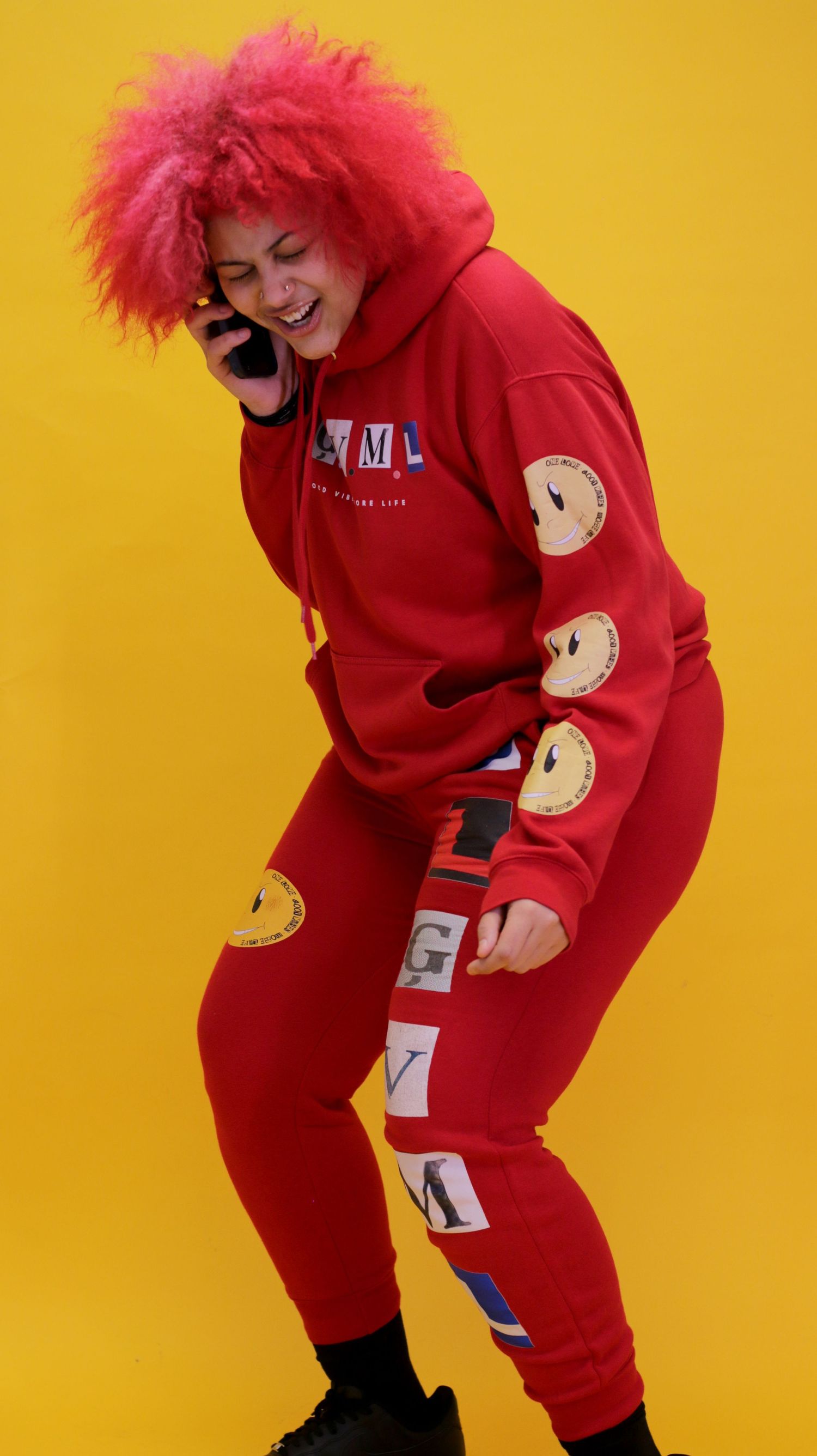 Person in red OLGVML tracksuit with pink afro wig laughs while talking on phone.