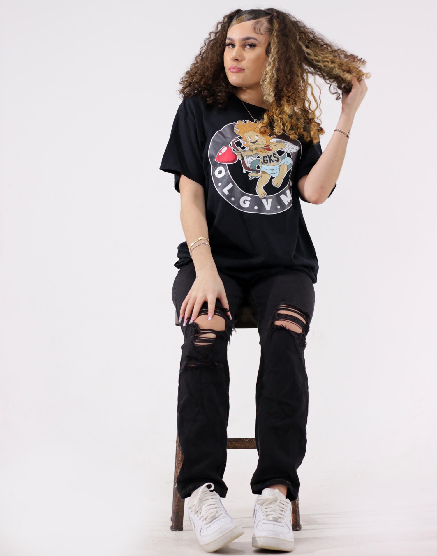 Woman in black OGVM graphic tee and ripped jeans sitting on stool in white studio.