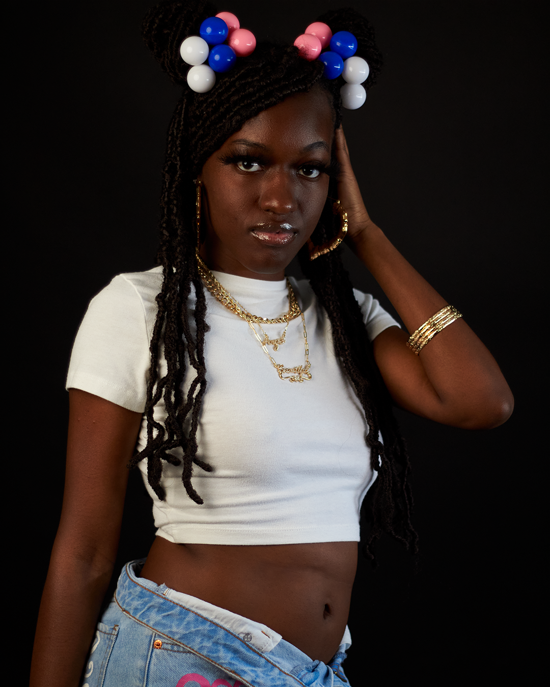Young Black woman with braided hair and colorful pom-poms in white crop top with gold jewelry posing.