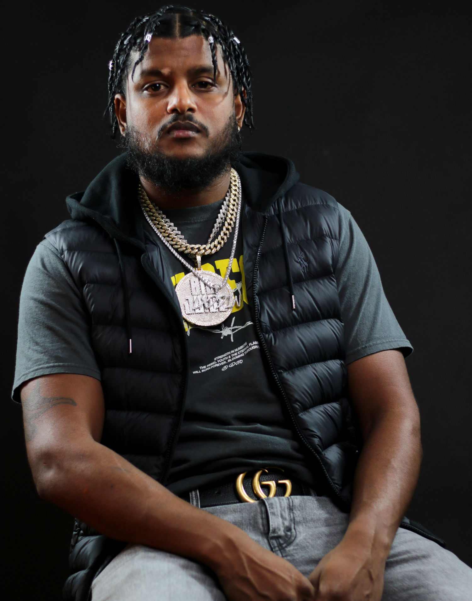 Bearded man in black puffer vest and gold chains sits confidently against a dark studio background.