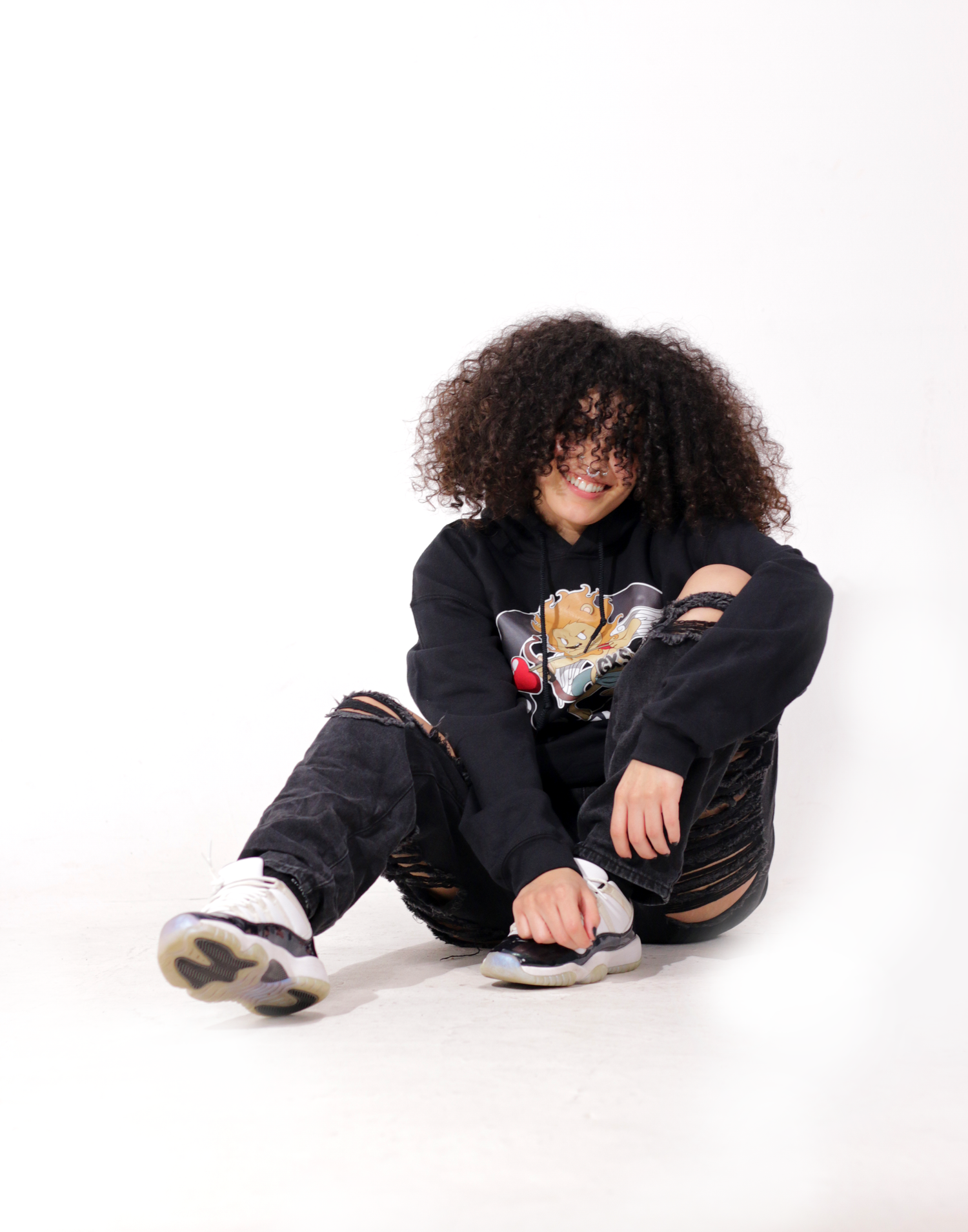 Person with curly hair sitting on floor in black OGVM hoodie and ripped jeans with sneakers.