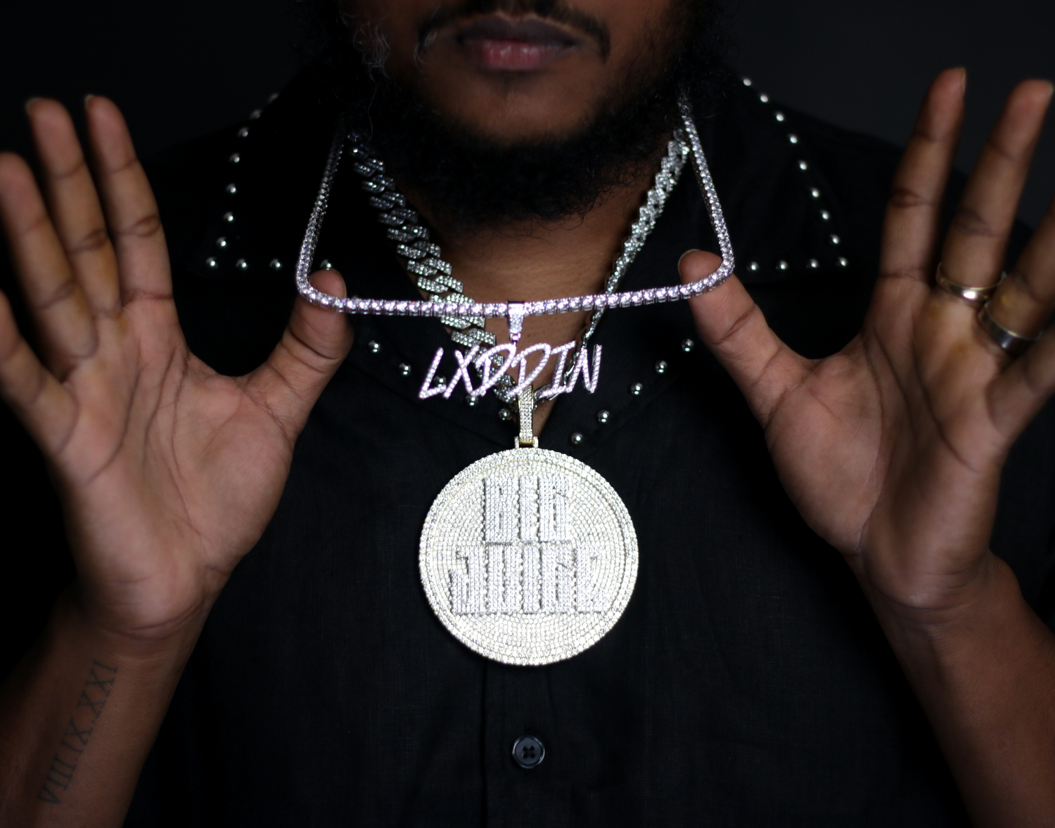 Close-up of man displaying diamond pendant necklace and chain with LXDDIN nameplate over black shirt.