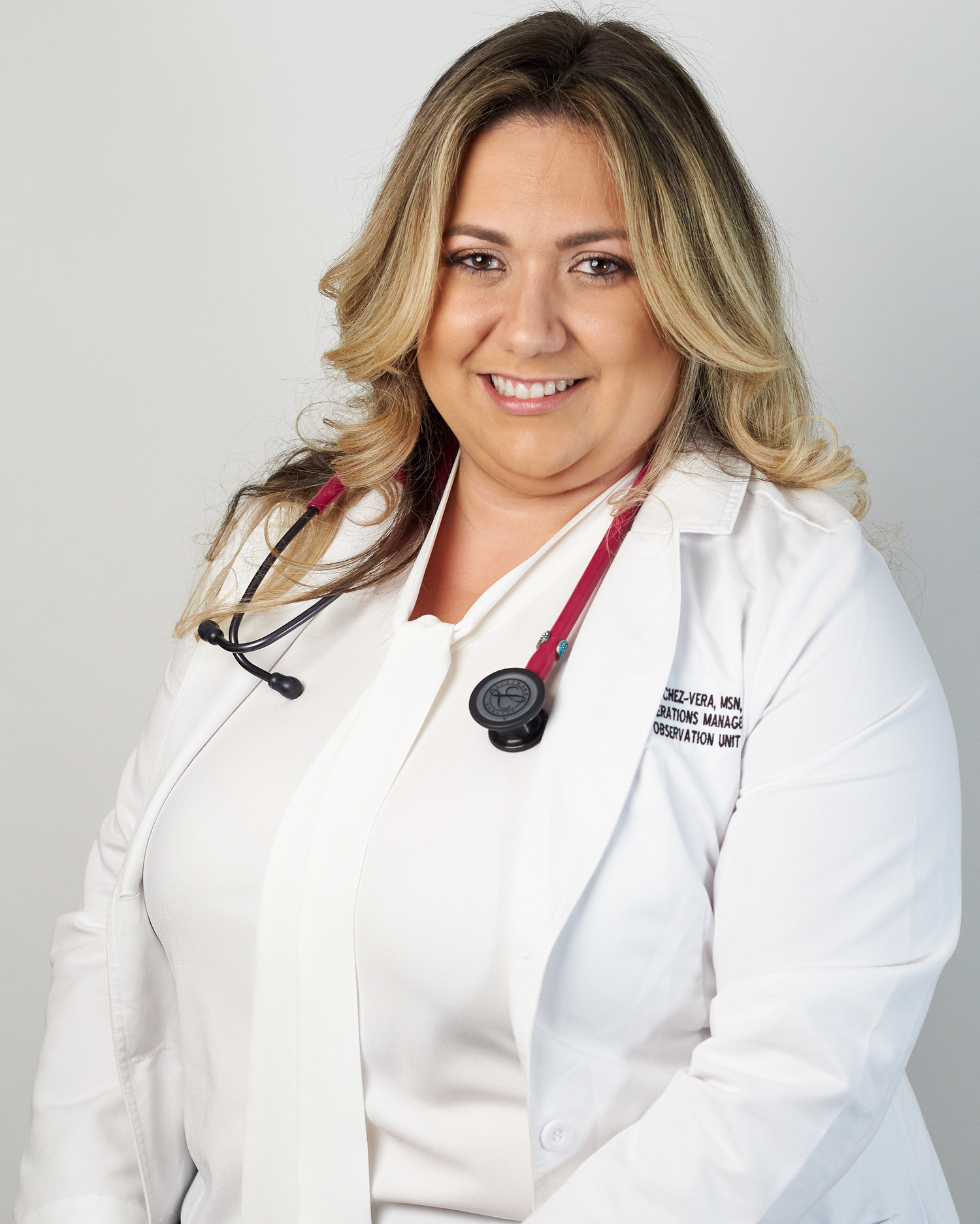 Female healthcare professional in white lab coat with red stethoscope poses for professional headshot.
