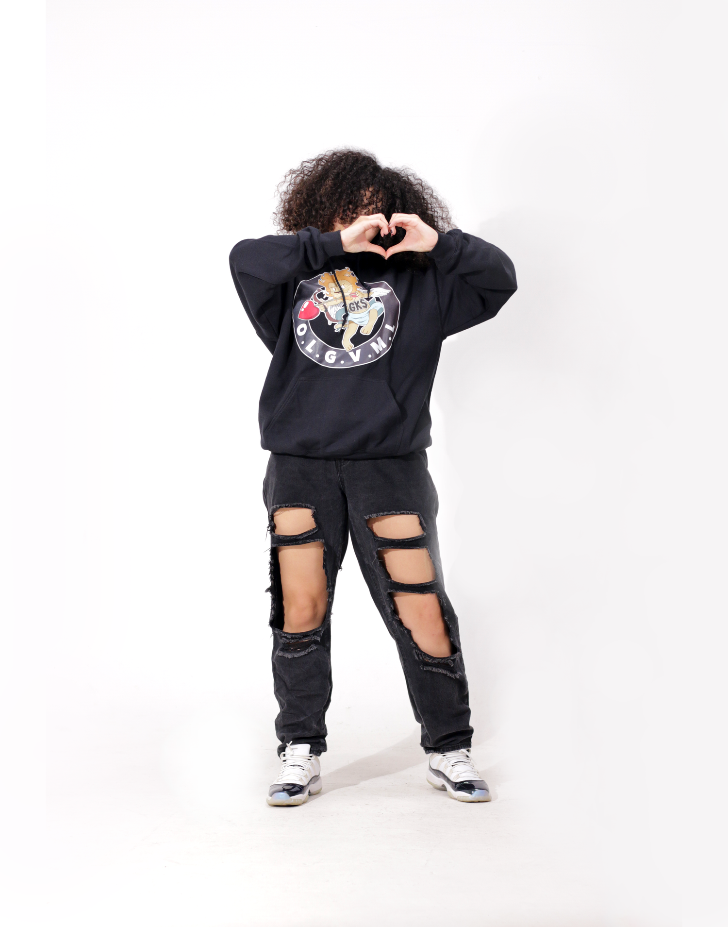 Person in black OGVM hoodie and ripped jeans making heart shape with hands over face.