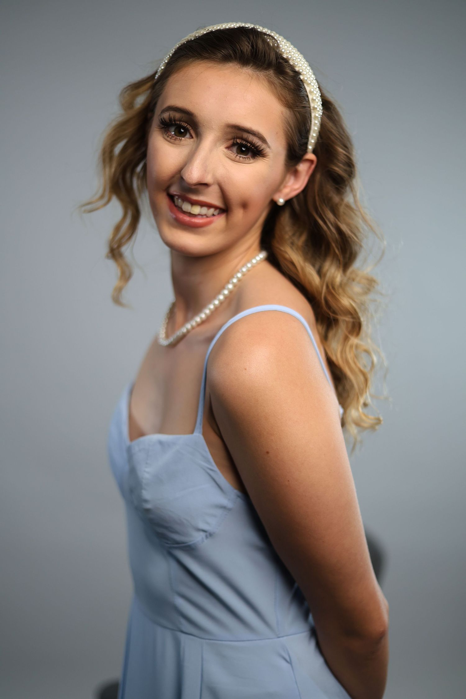 Smiling young woman in light blue dress with pearl necklace and headband on gray background.