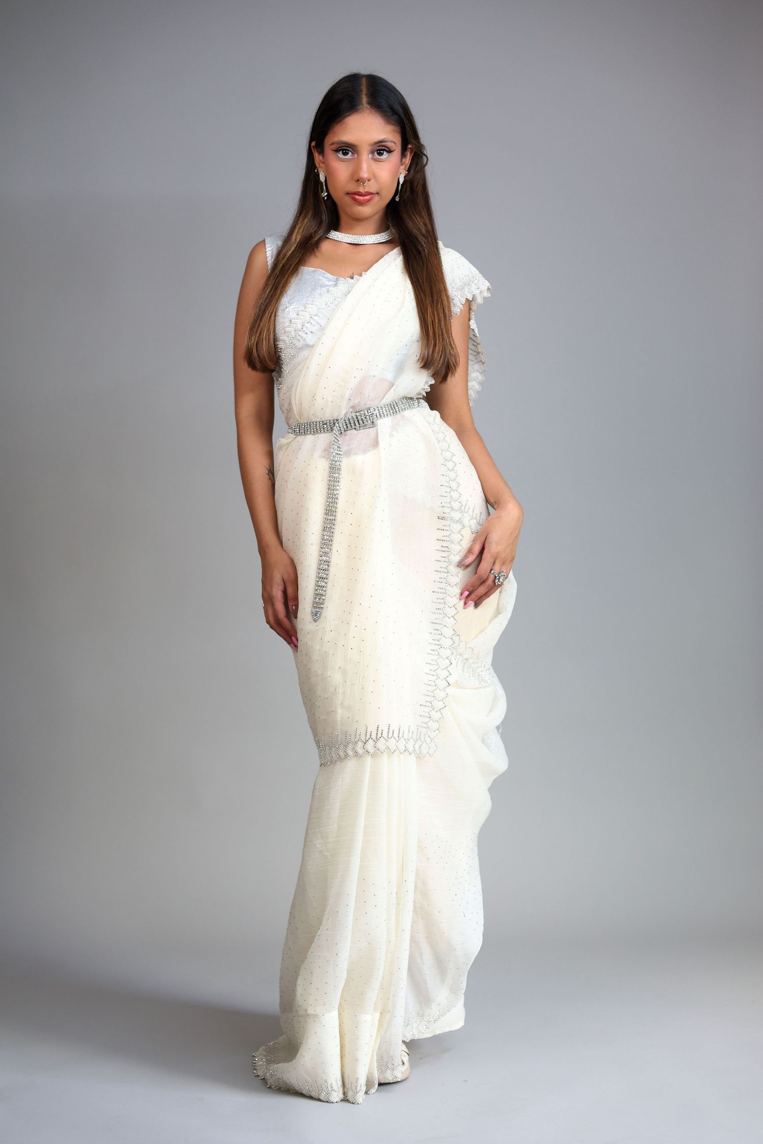 Young woman poses elegantly in white embroidered saree with silver belt and choker necklace in studio.