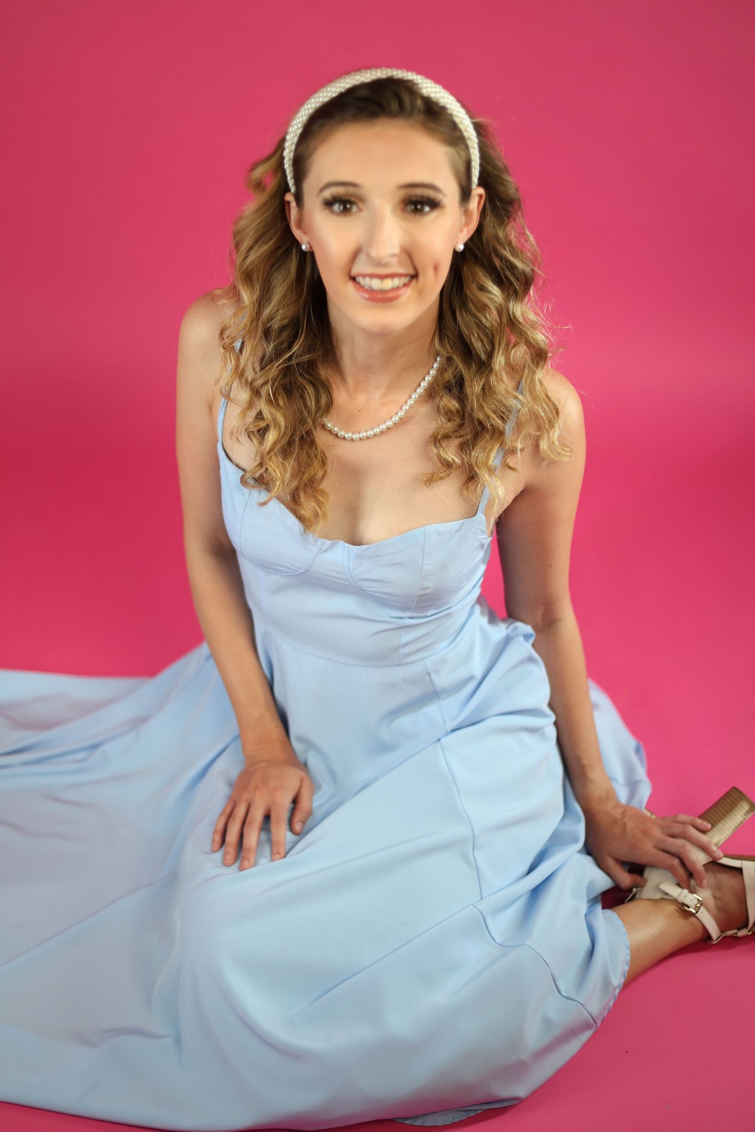 Young woman in light blue prom dress and pearl necklace sitting on pink background smiling.