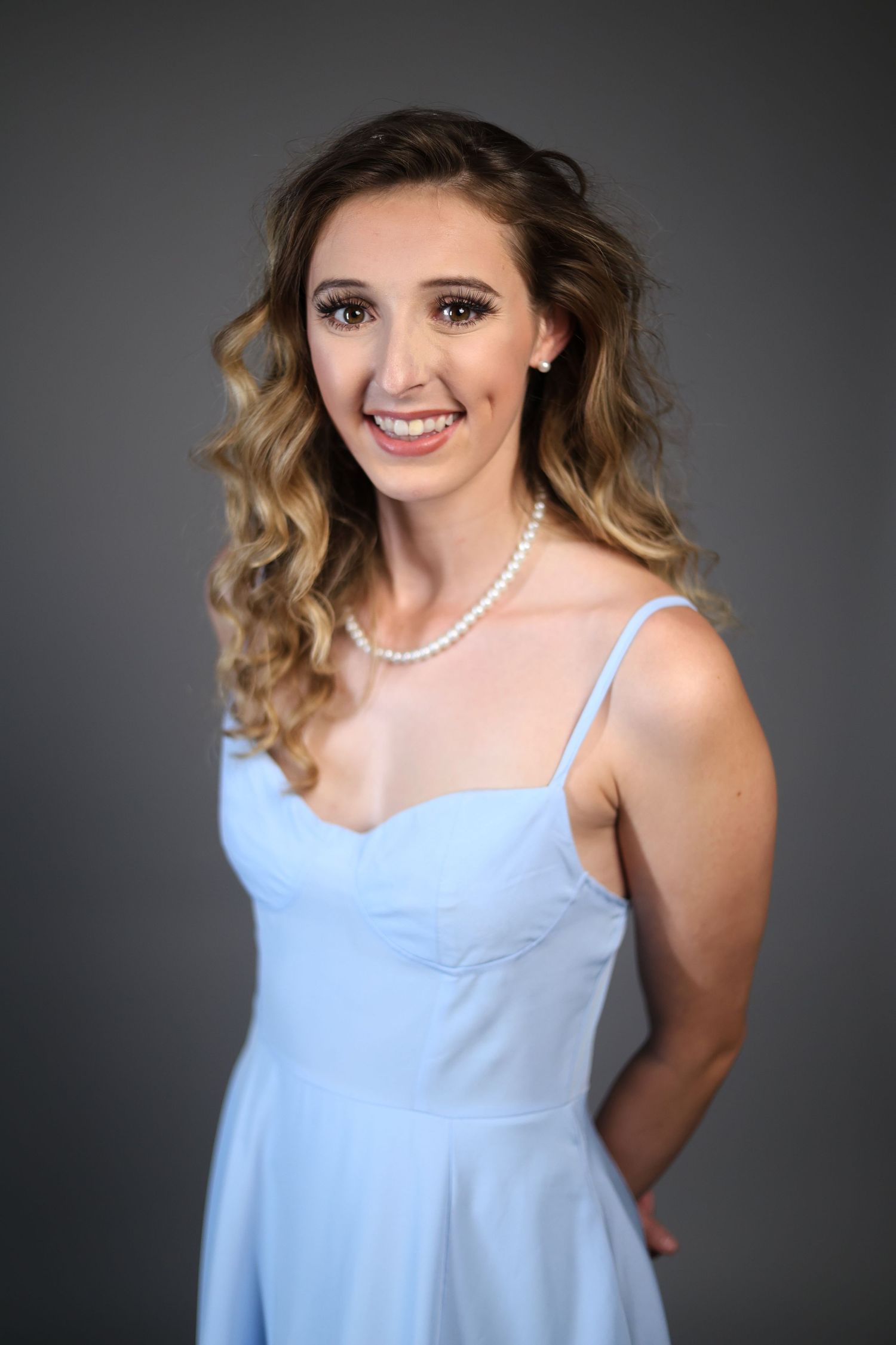 Young woman in baby blue sundress and pearl necklace posing against gray studio backdrop.