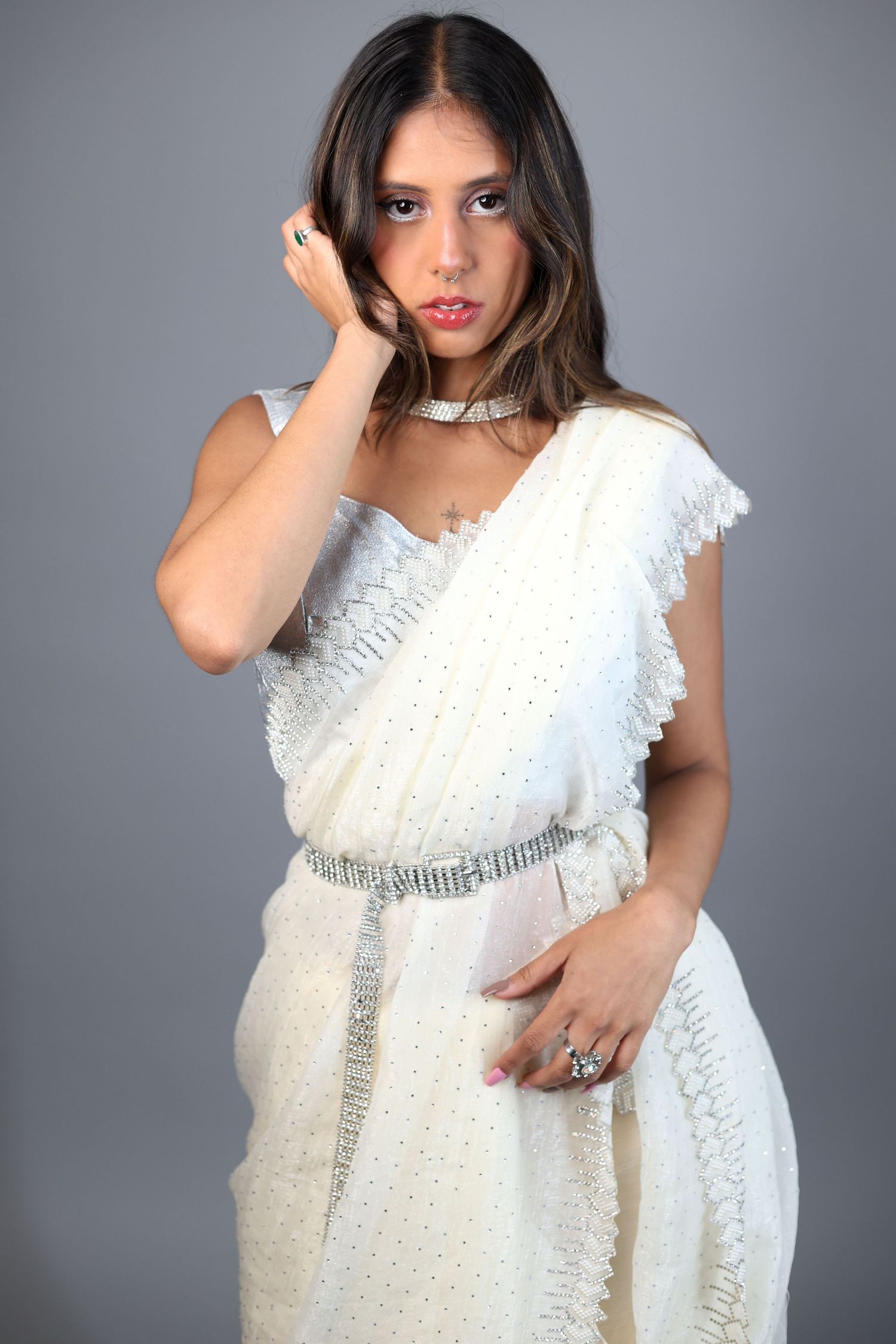 Young woman in white saree with silver choker and belt posing with hand in hair against gray background.