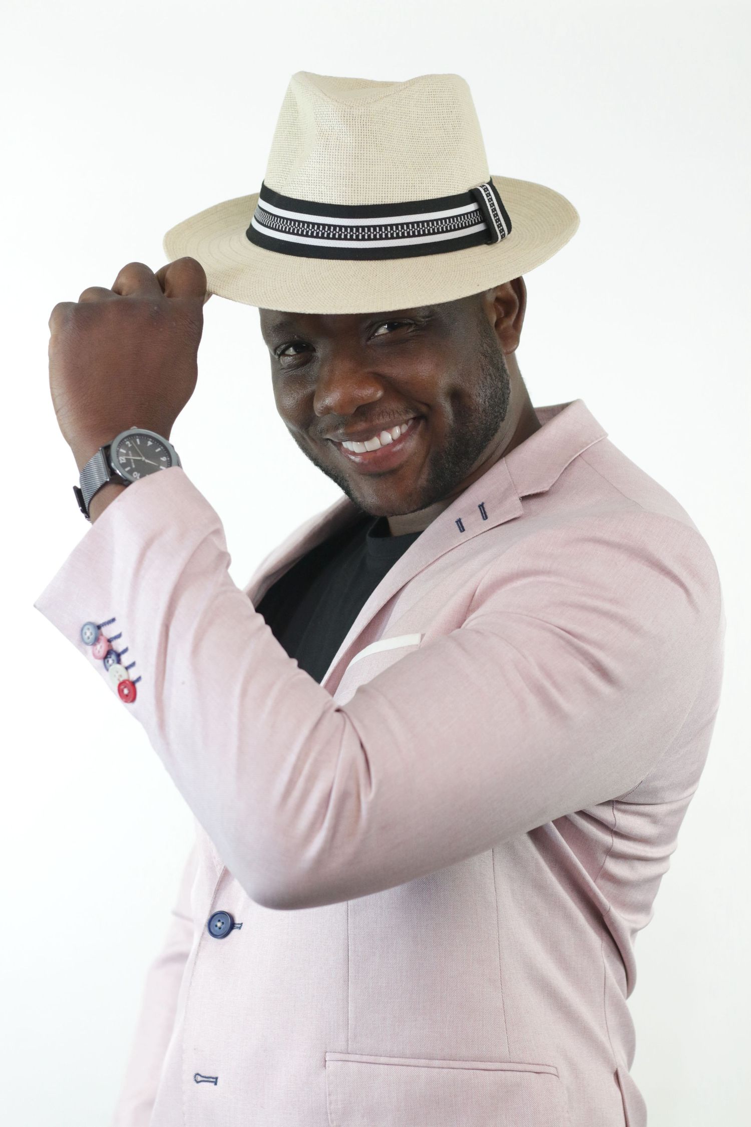 Smiling man in pink blazer tipping cream fedora hat against white studio background.