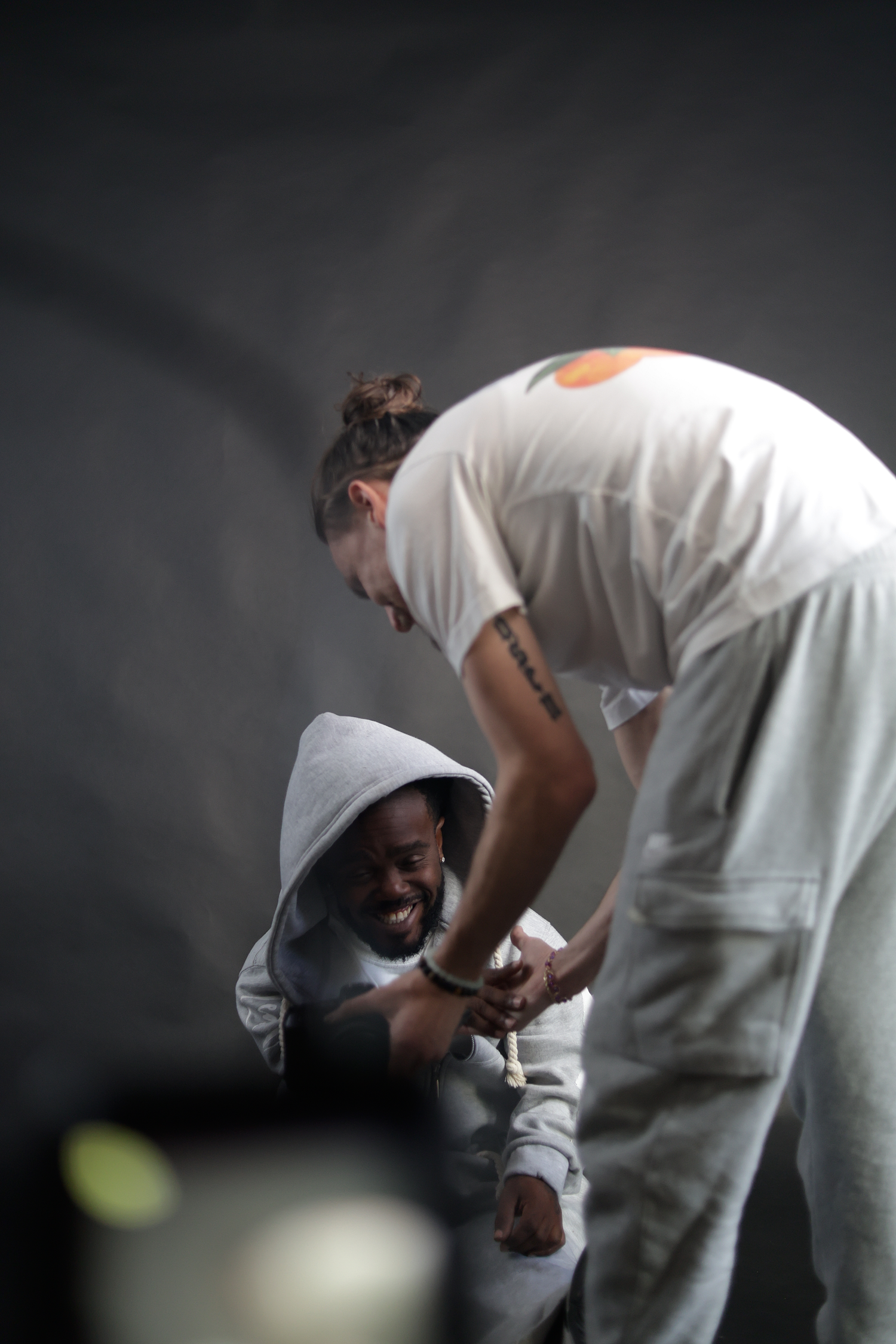 Two men interact playfully in a studio, one in a hoodie and one in white overalls against a dark backdrop.