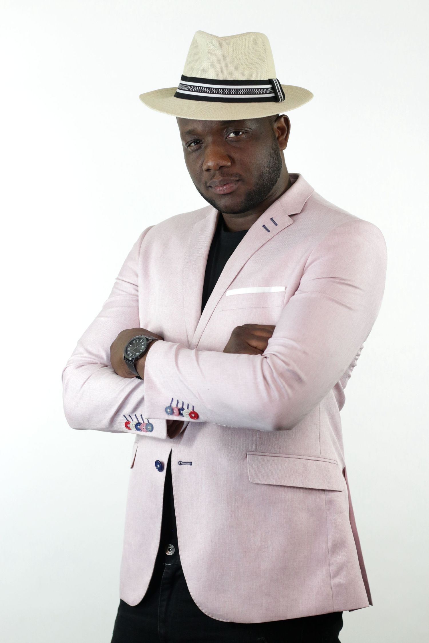 Stylish man in pink blazer and cream fedora hat posing with arms crossed against white background.