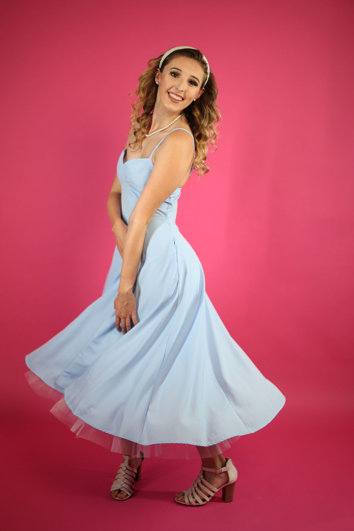 Young woman in light blue midi dress twirling on pink background wearing strappy heels.