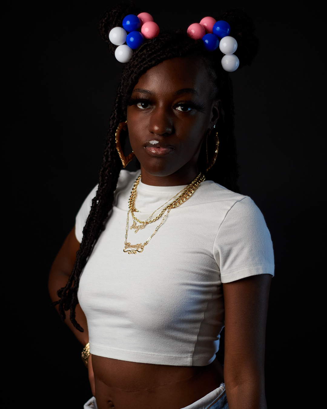 Young Black woman with braided hair and colorful pom-poms wearing white crop top and gold chain necklaces.