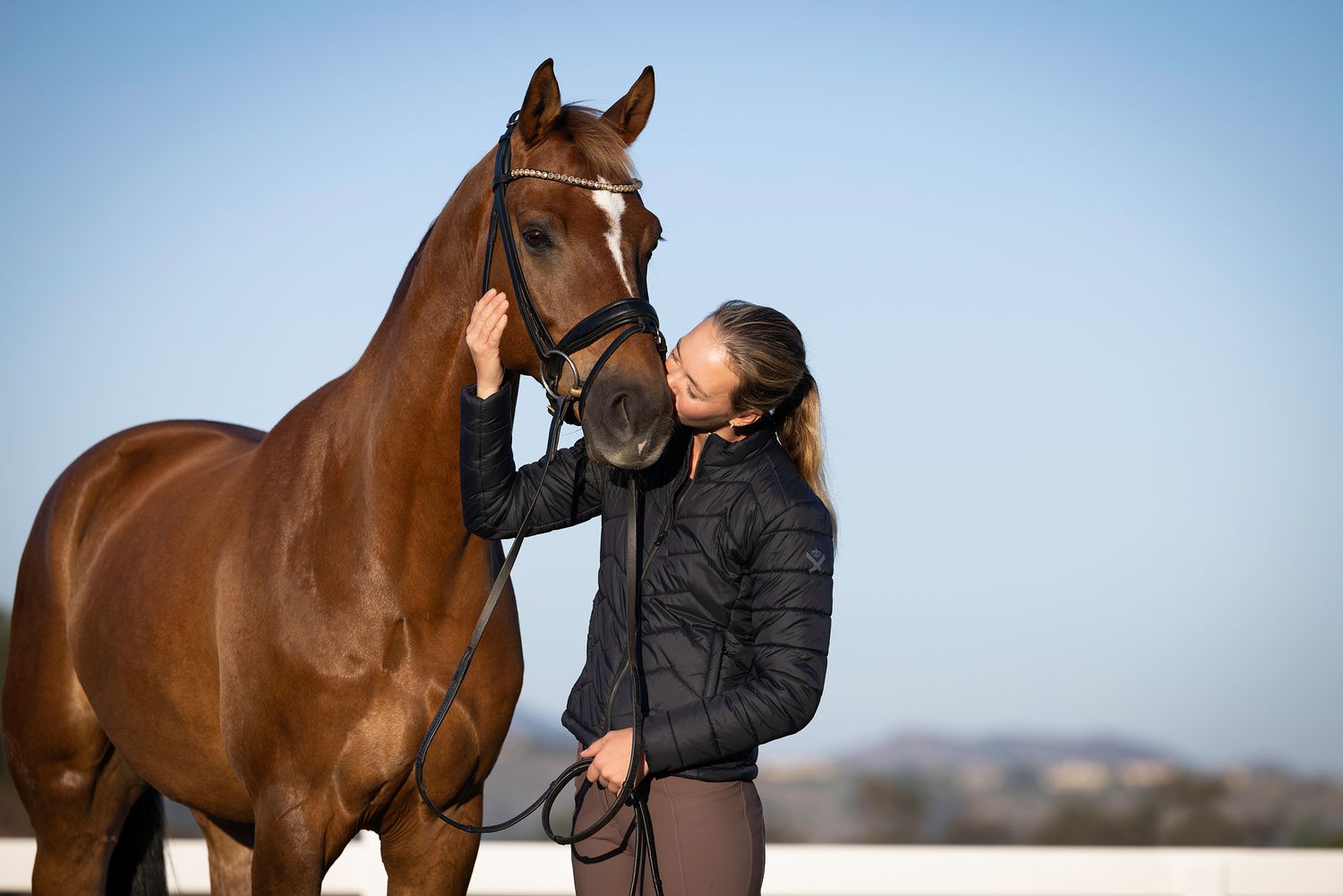 Woman in a black quilted jacket gently kisses the nose of a chestnut horse with a white blaze against a clear blue sky.