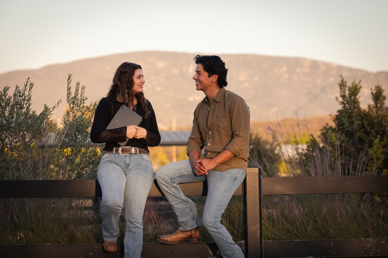 A smiling young woman holding a tablet and a man in a khaki shirt chat while sitting on a wooden fence with mountains behind them.
