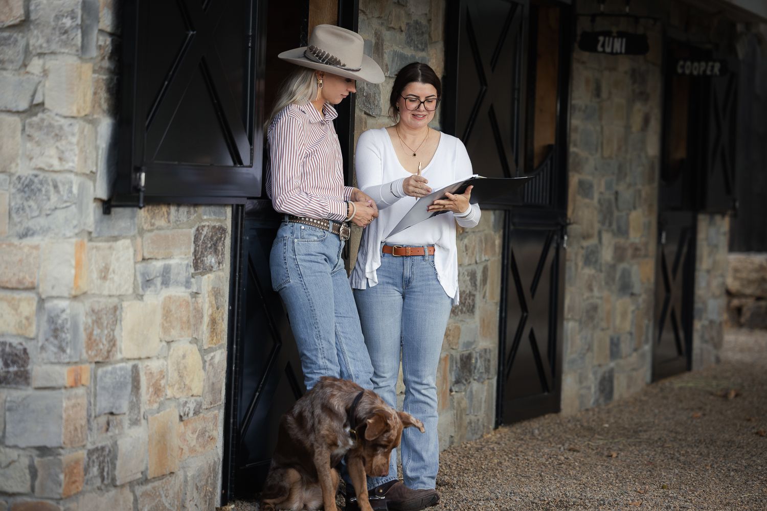 Two women in jeans, one in a cowboy hat, chat beside a stone stable with black doors while a brown dog sits at their feet.