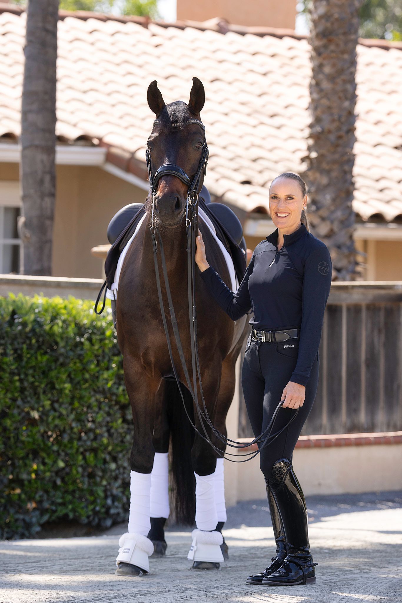Smiling woman in black riding attire stands beside a bay dressage horse wearing white leg wraps and a saddle outside a tile-roofed stable.