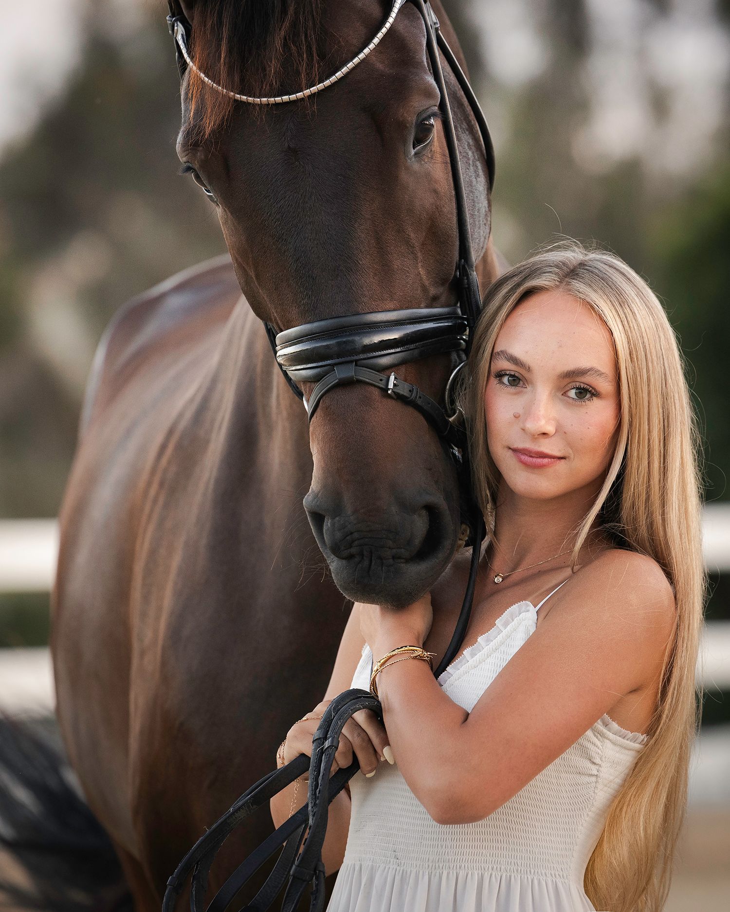 Young woman with long blonde hair in a white sundress stands close to a bay horse wearing a black leather bridle outdoors.