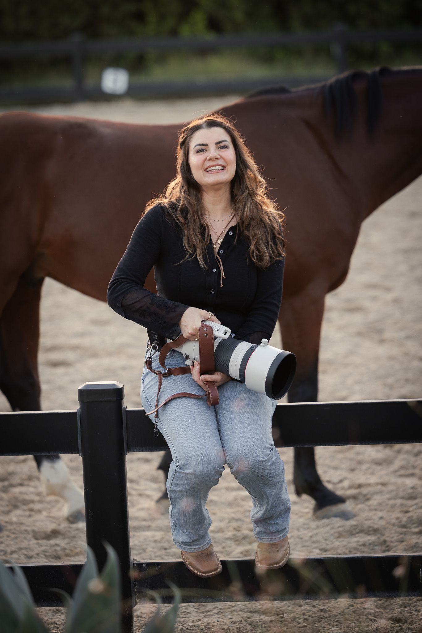 Smiling woman sits on a wooden arena fence holding a camera with a telephoto lens, with a bay horse standing behind her on sandy ground.