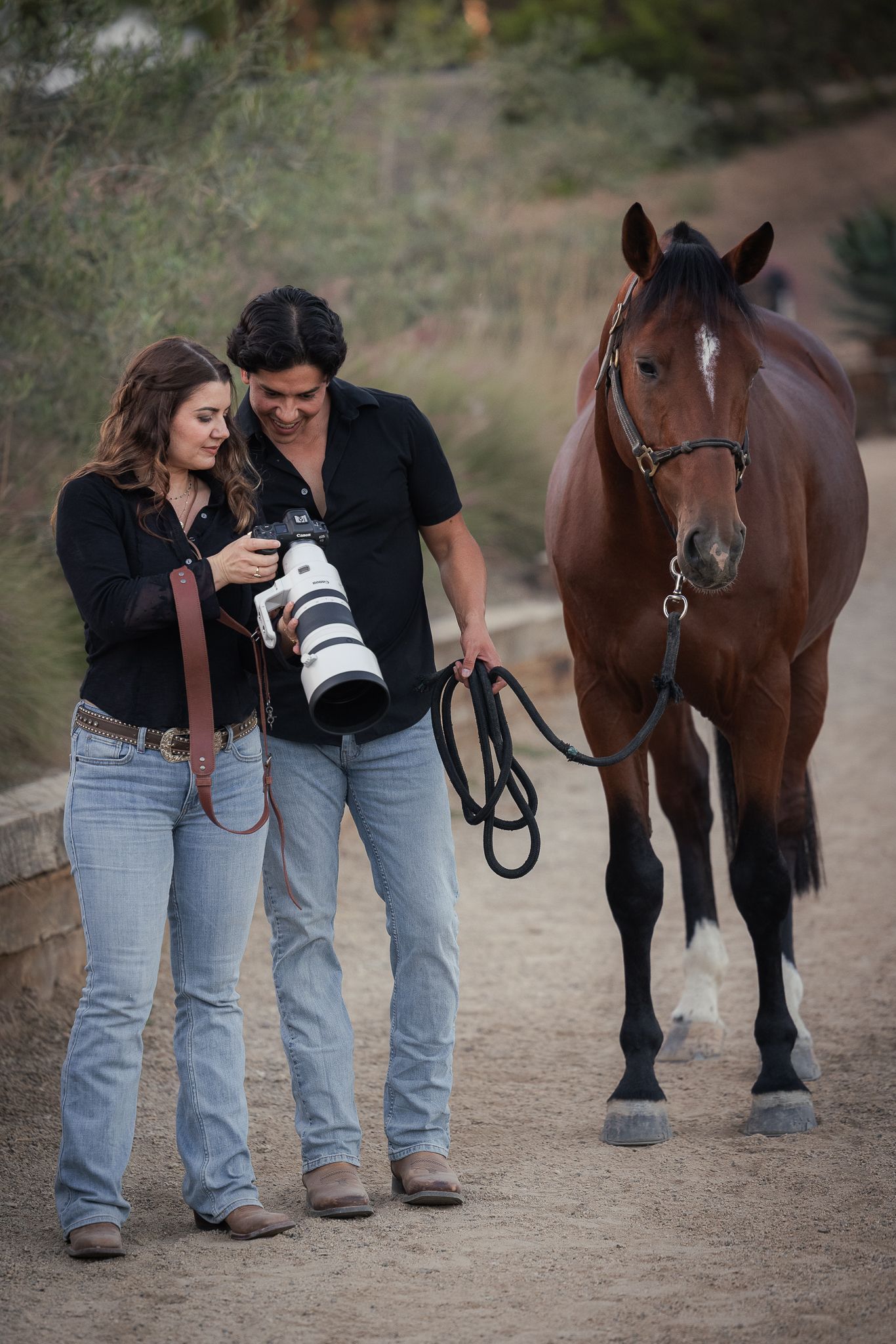 Photographer and a man in jeans review shots on a camera with a telephoto lens while leading a bay horse along a dirt path.