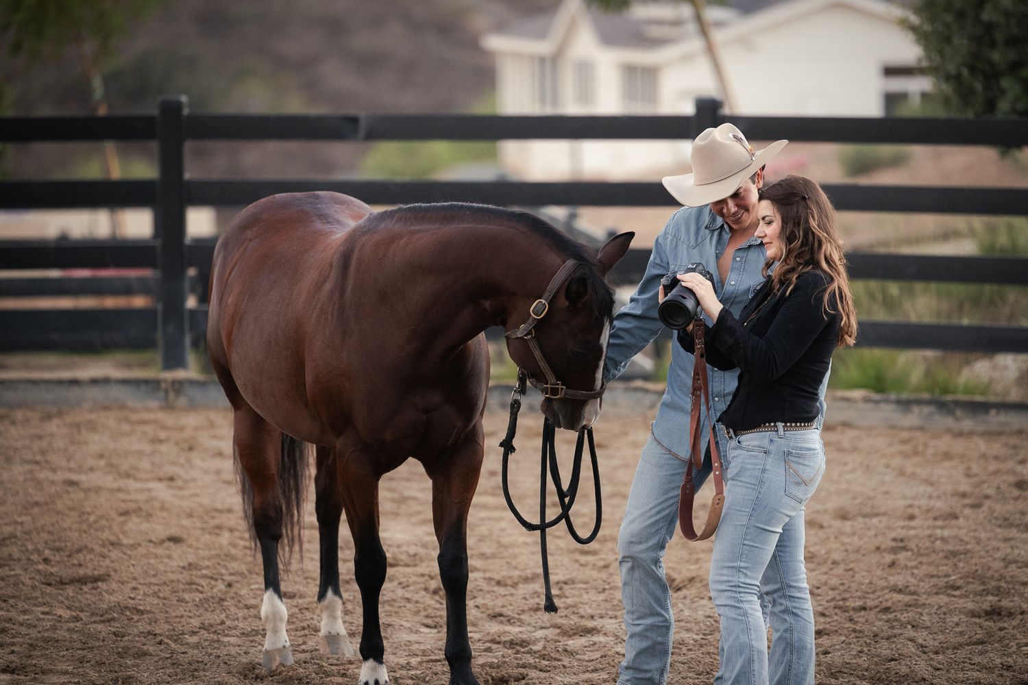 Two people in cowboy hats stand beside a bay horse in a dirt riding arena, holding its lead rope near a wooden ranch fence.