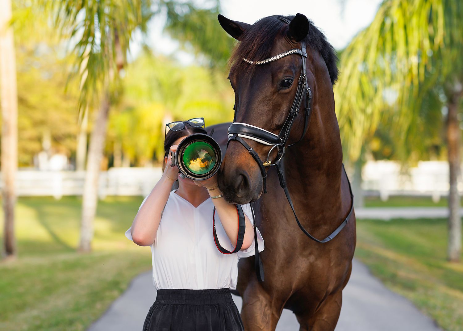 Photographer in white blouse and black skirt aiming a large telephoto lens at a brown bridled horse on a palm-lined path.