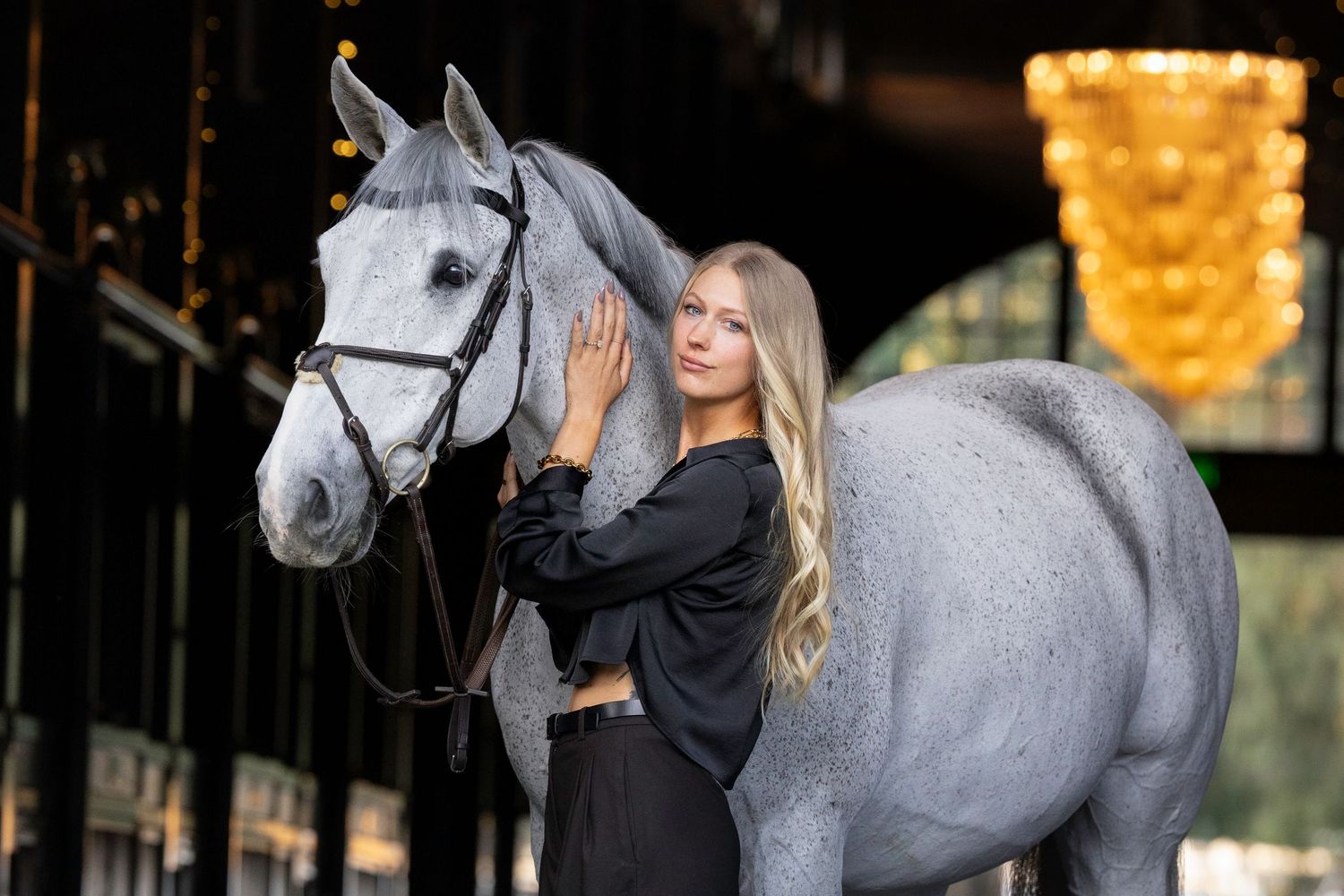 Blonde woman in a black top and pants stands beside a dapple gray horse wearing a bridle in an elegant stable with a golden chandelier.