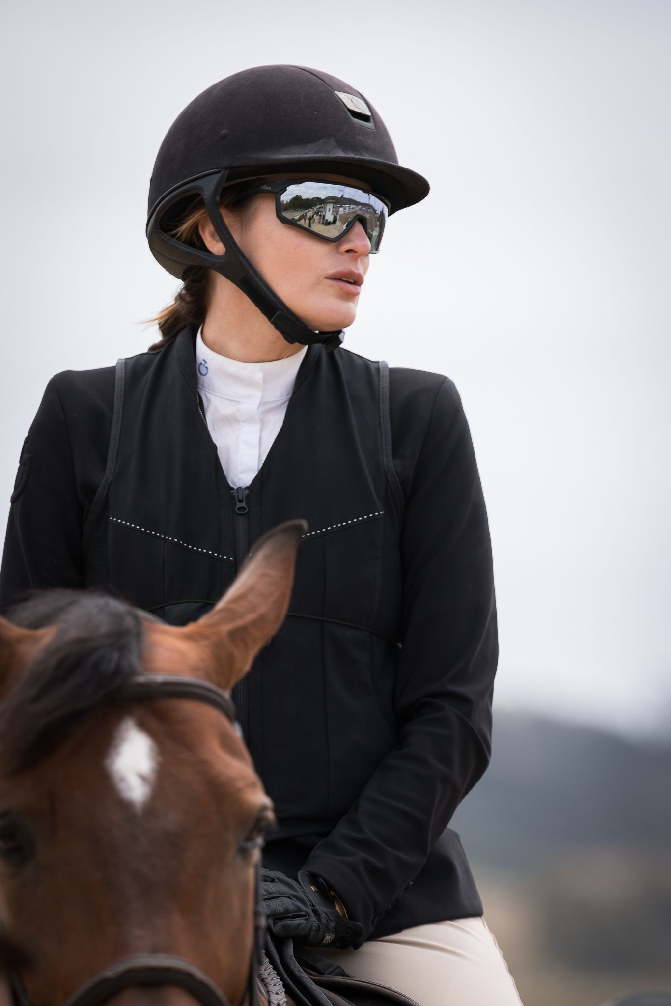 Female equestrian rider in a black helmet, mirrored sport sunglasses and fitted black riding jacket sitting on a brown horse outdoors.