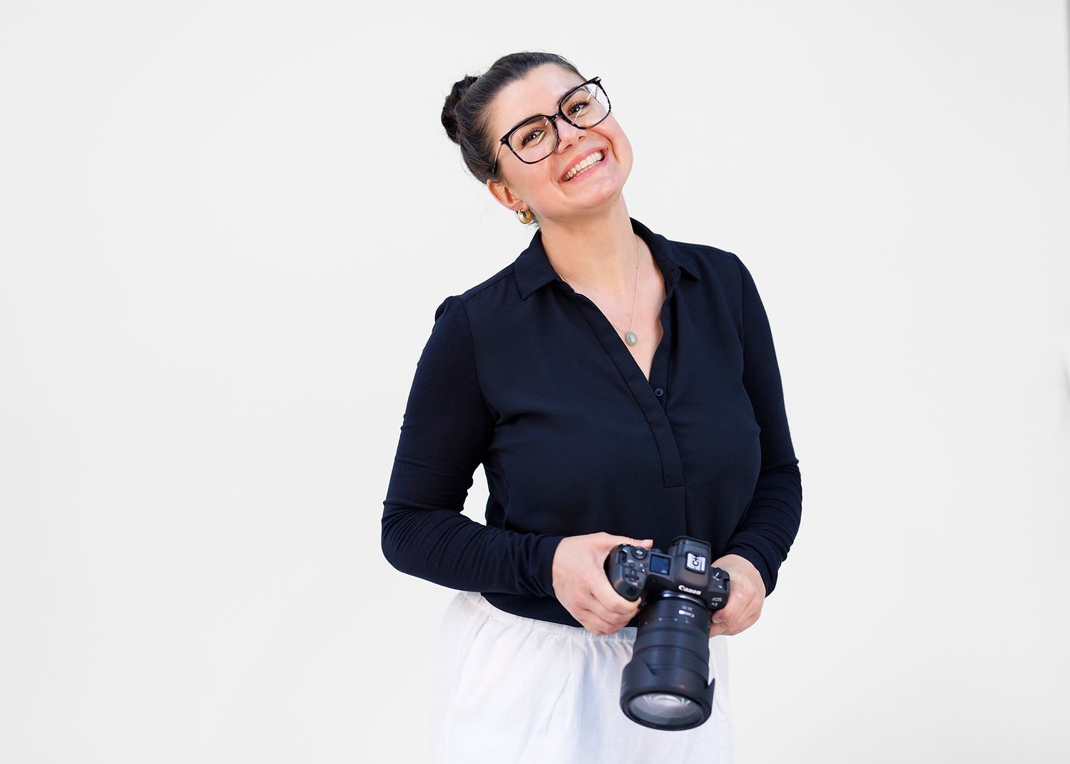 Smiling woman with glasses and dark hair in a bun holds a black DSLR camera while posing in a navy blouse against a white background.
