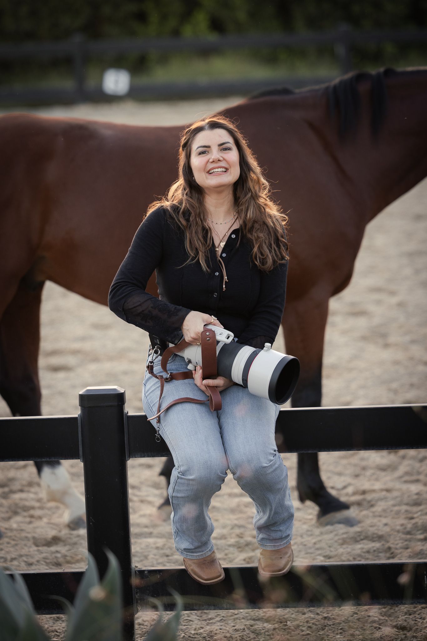 Smiling woman with long wavy hair sits on a black fence holding a camera with a telephoto lens, a brown horse behind her in a sandy arena.