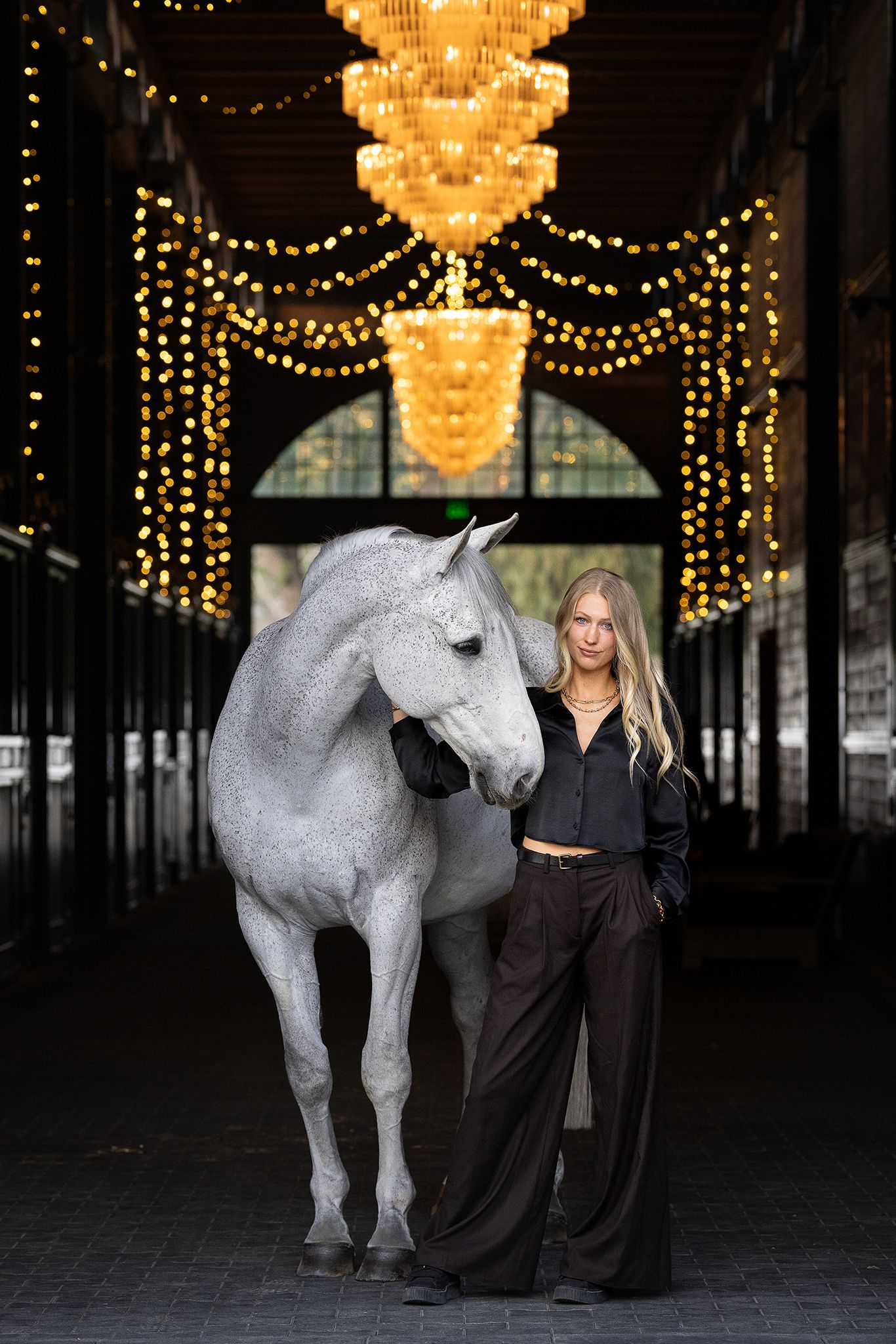 Woman in black blouse and wide-leg trousers stands beside a gray horse in a stable aisle lit by golden chandeliers and string lights.