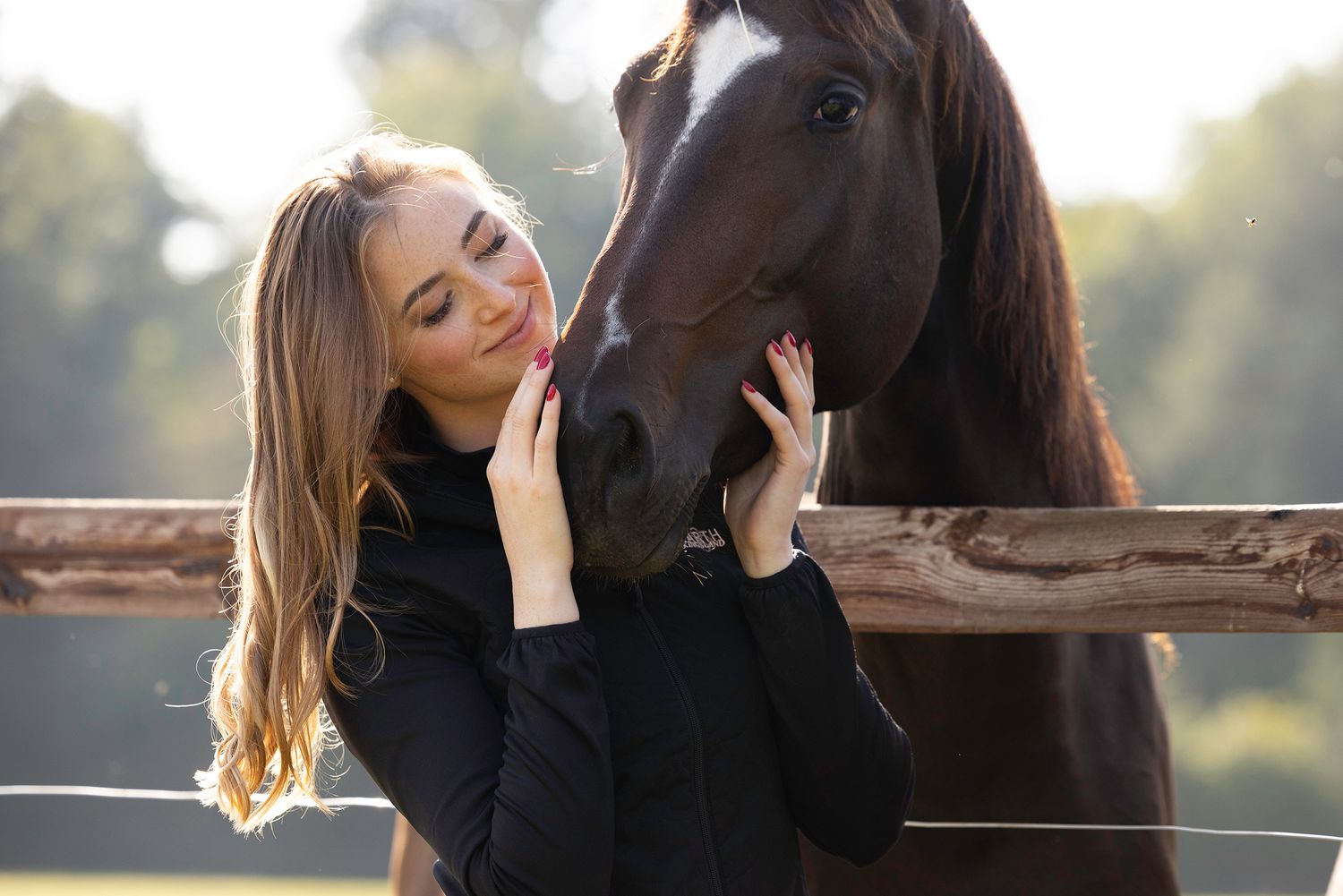 Smiling young woman with long blonde hair gently touches the face of a dark brown horse beside a wooden fence outdoors.