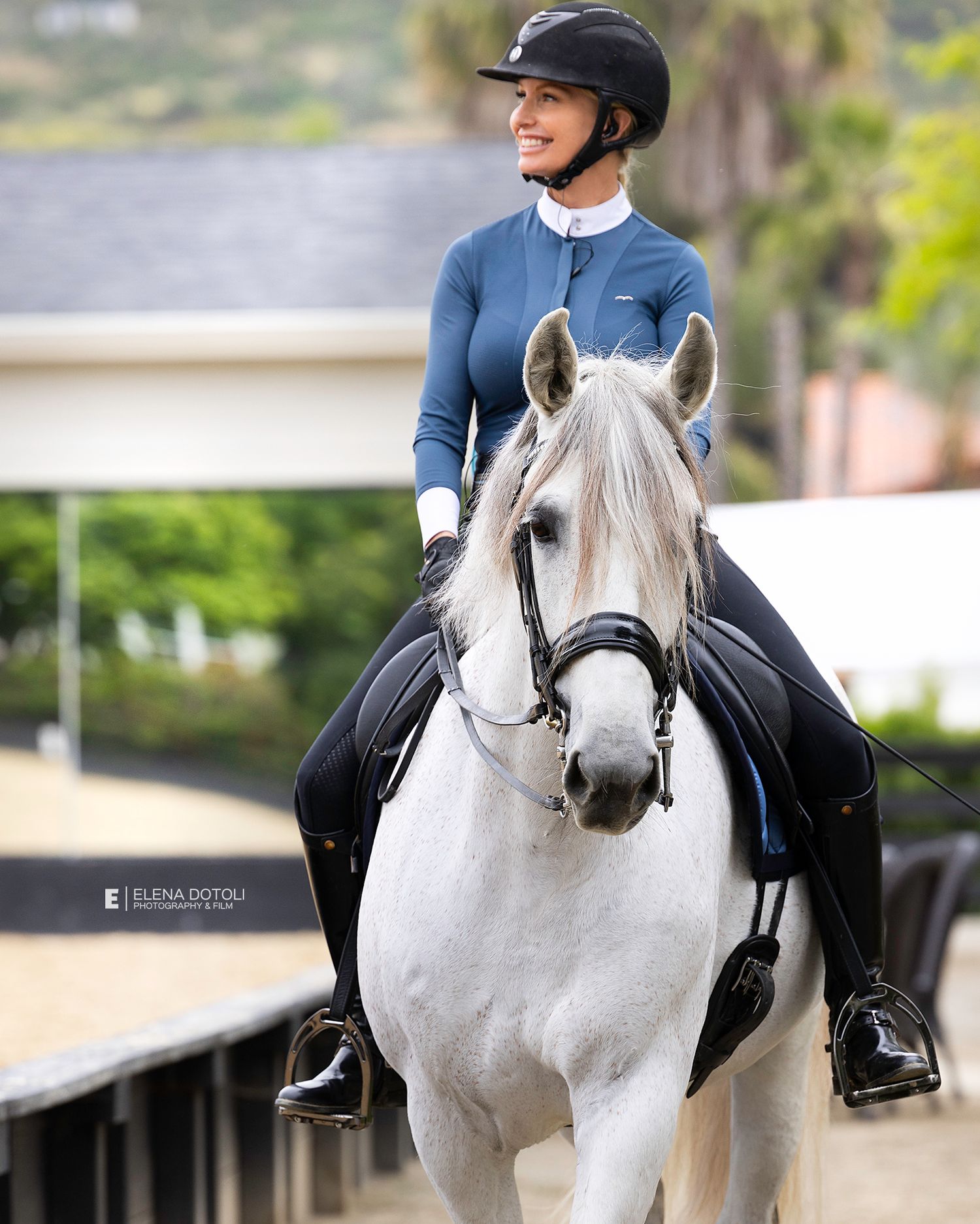 Smiling equestrian in a helmet and blue long-sleeve top rides a white horse in a bridle at a sunny outdoor dressage arena.