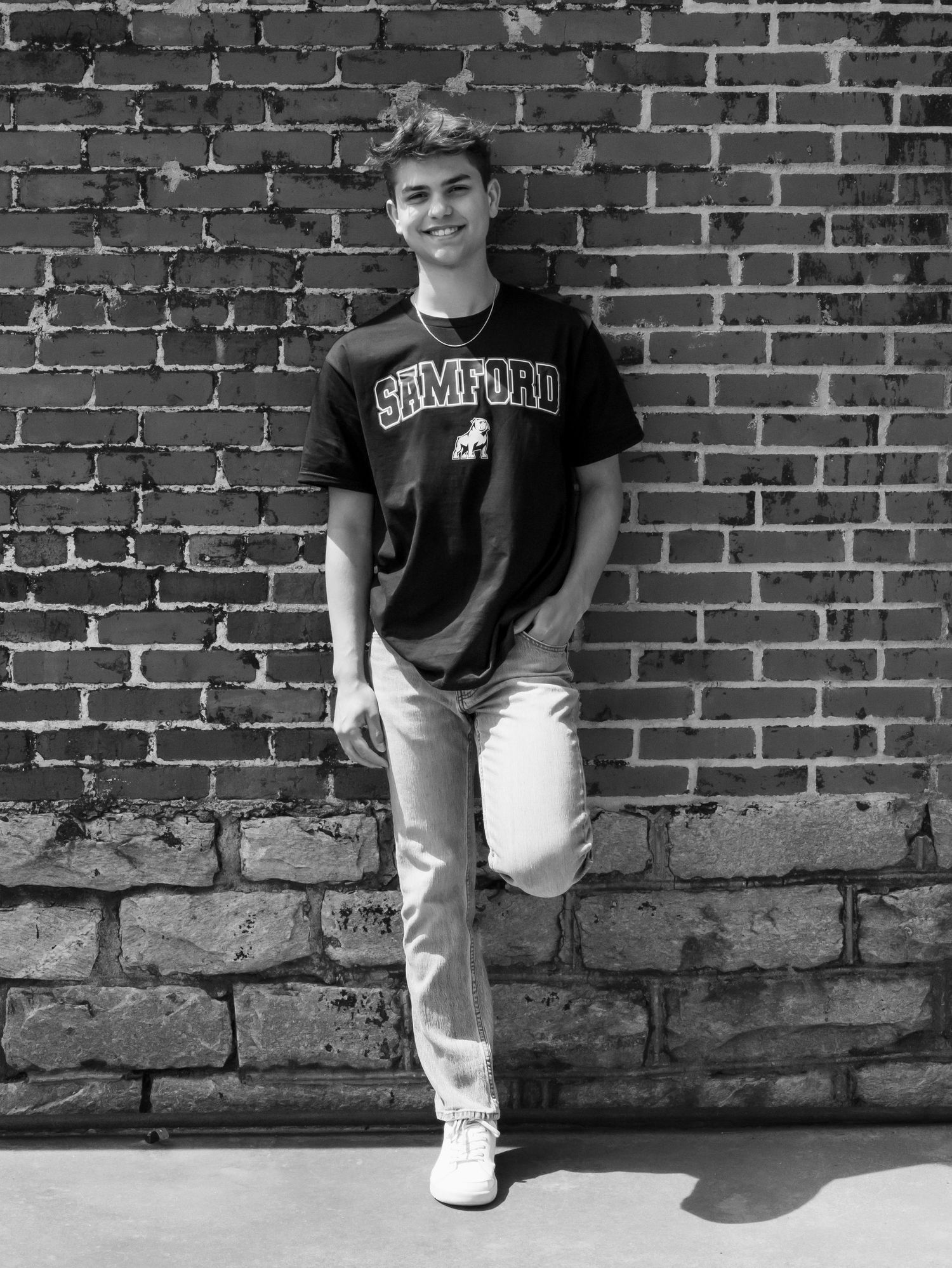 Young man in Samford University t-shirt leaning against brick wall, black and white senior portrait photo.