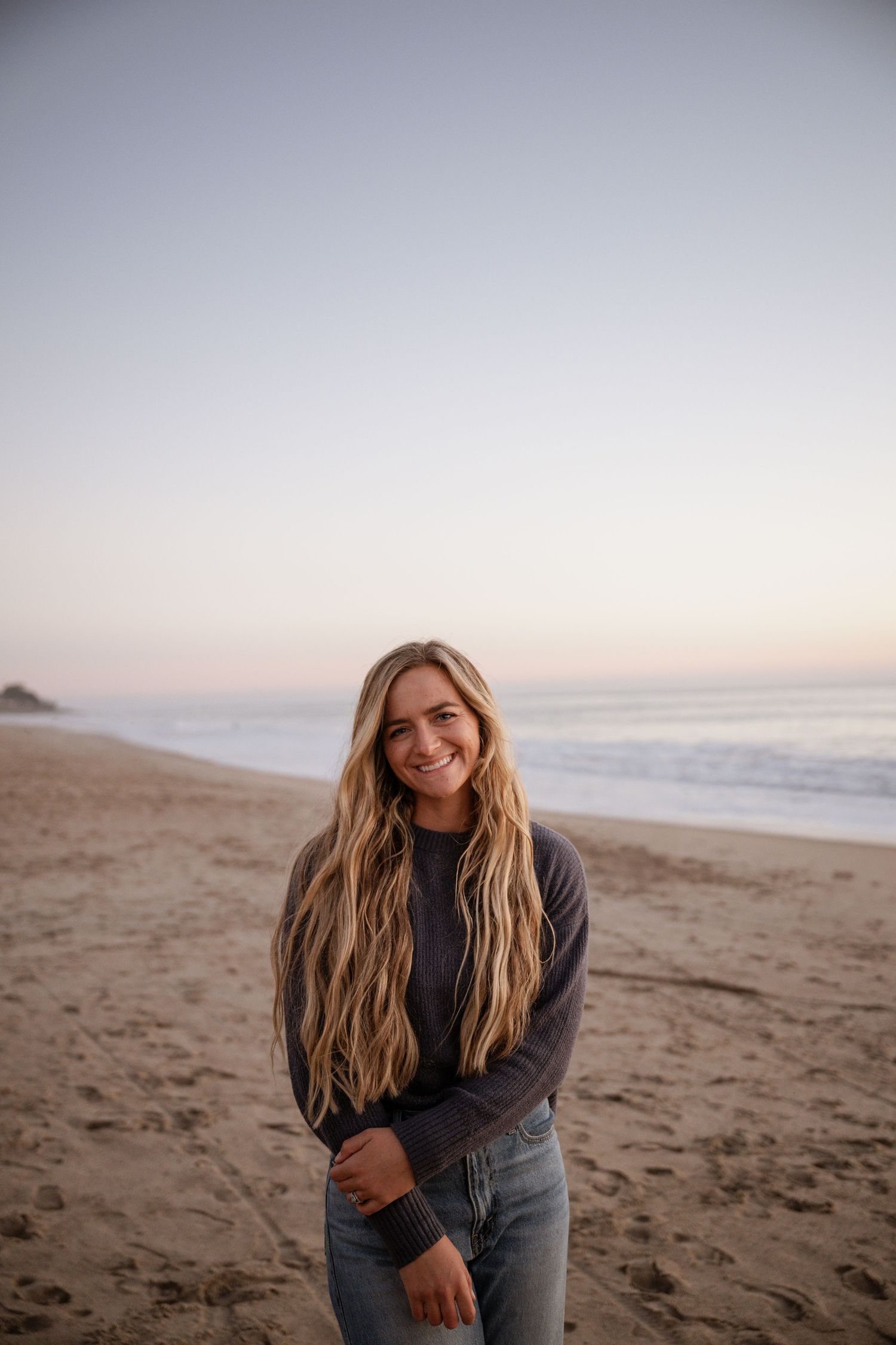 A person in jeans and a dark sweater sits casually on a sandy beach at sunset with long blonde hair flowing in the breeze.