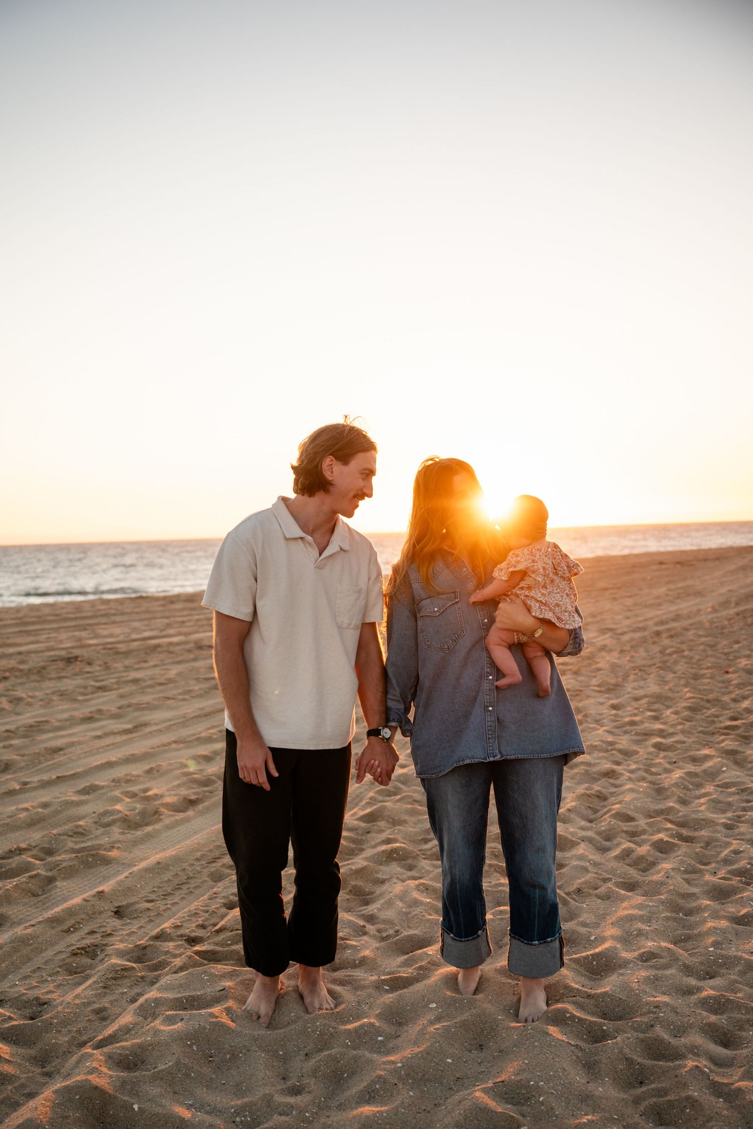 Two people walk barefoot along a sandy beach at sunset holding hands as golden sunlight illuminates the scene.