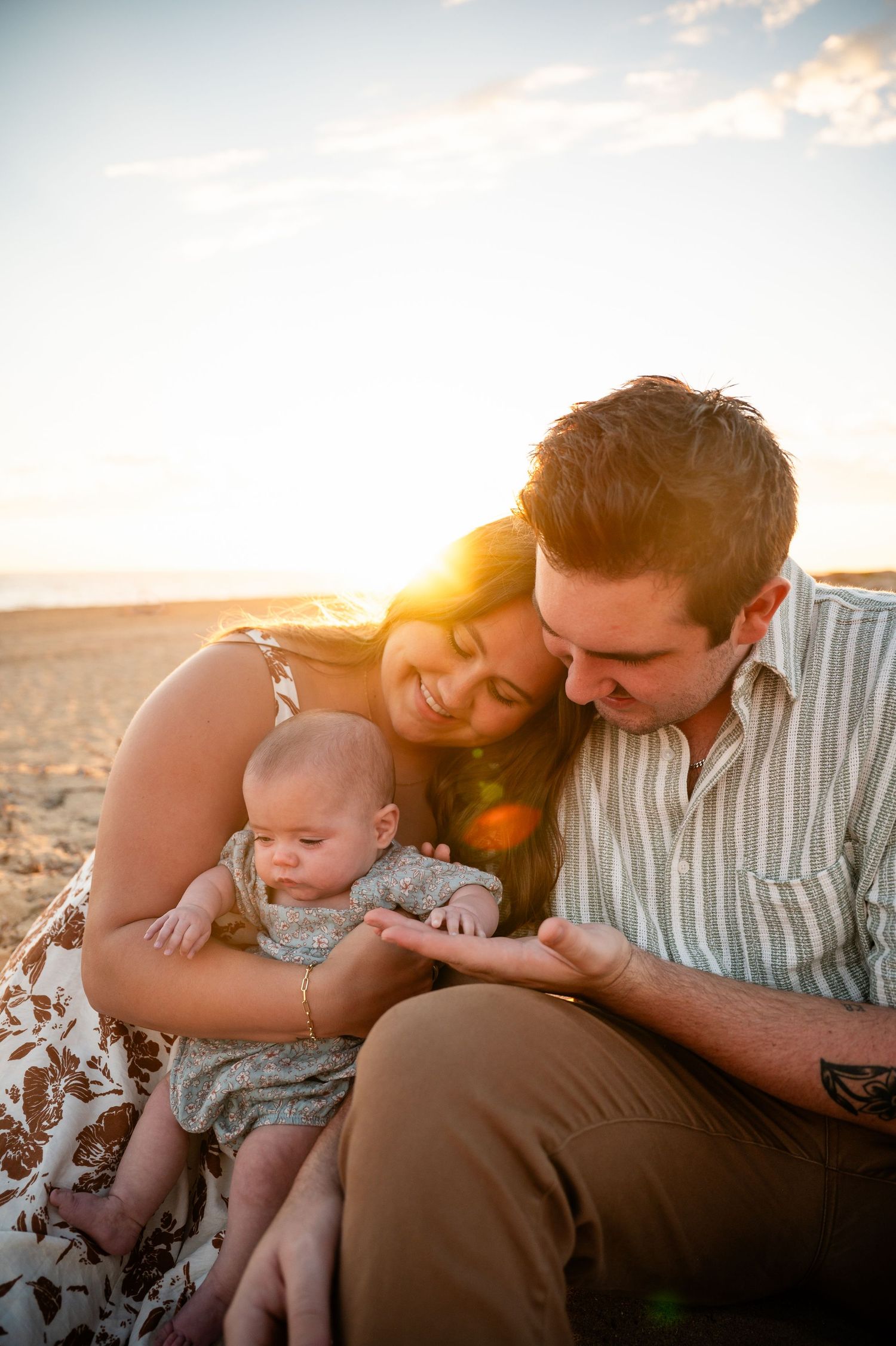 A family sits together on the beach at sunset with warm golden light and lens flare in the background.