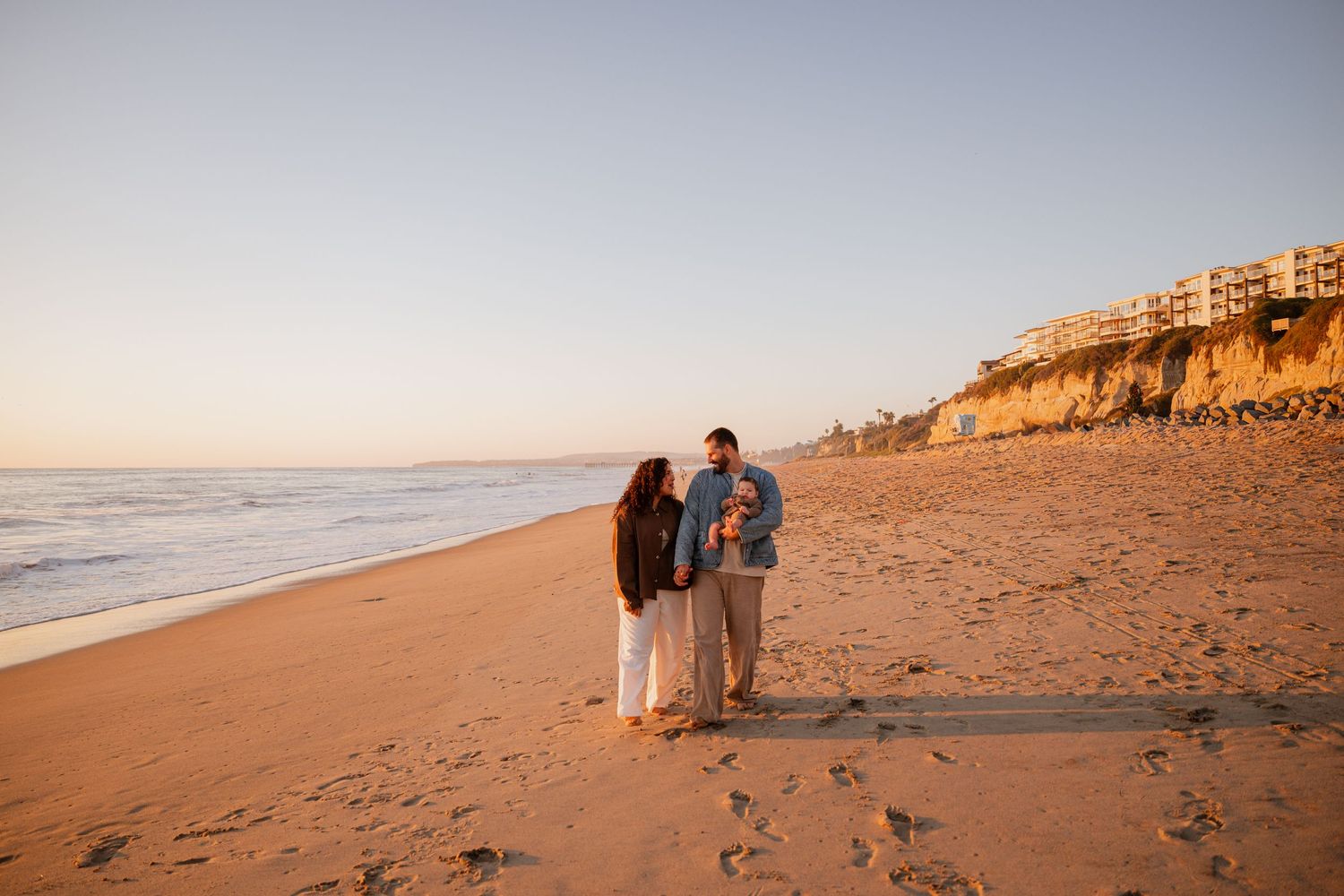 Romantic couple walking along a sandy beach at sunset with cliffs in the background.