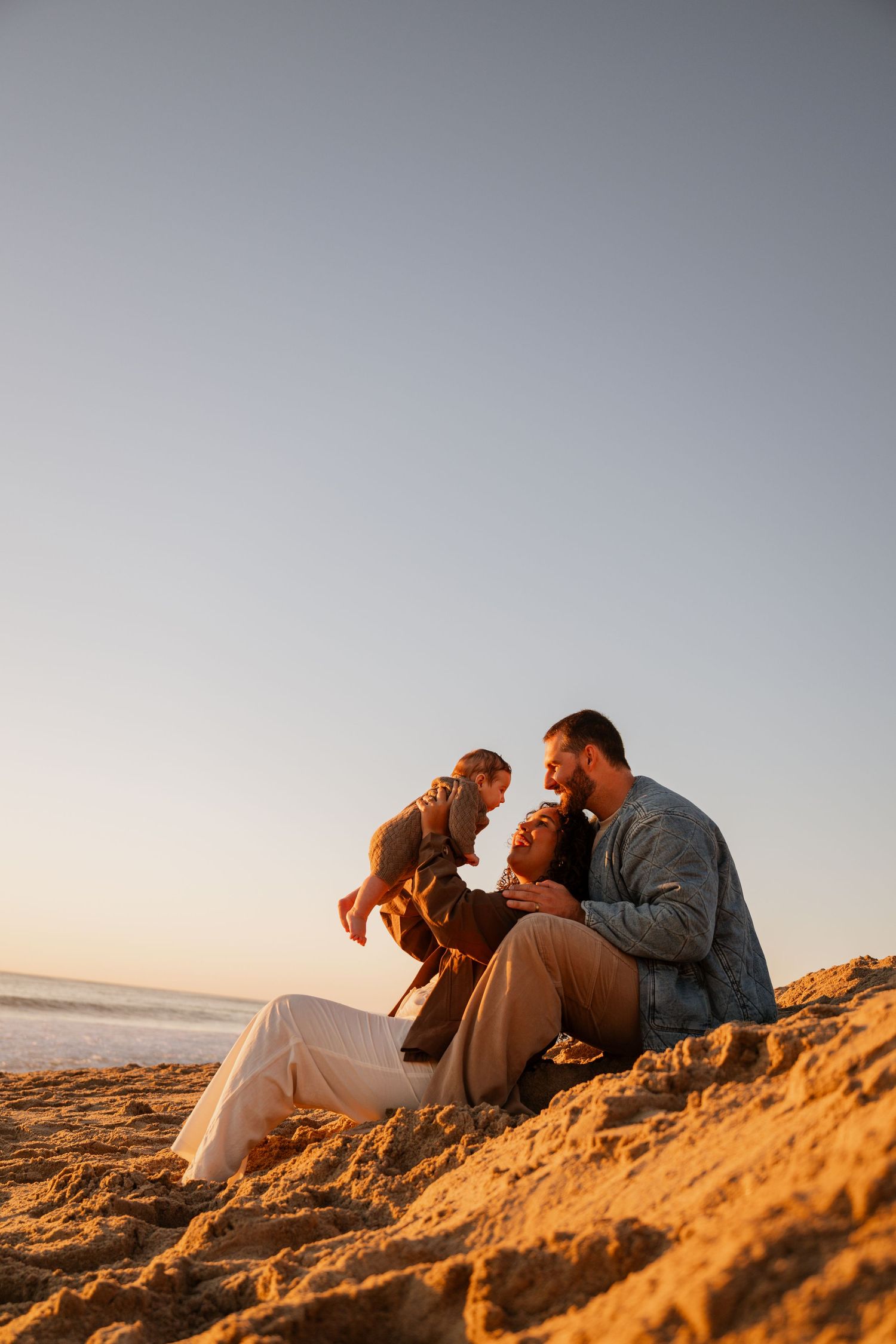 Couple sitting close together on sandy beach dunes during golden hour sunset by the ocean.