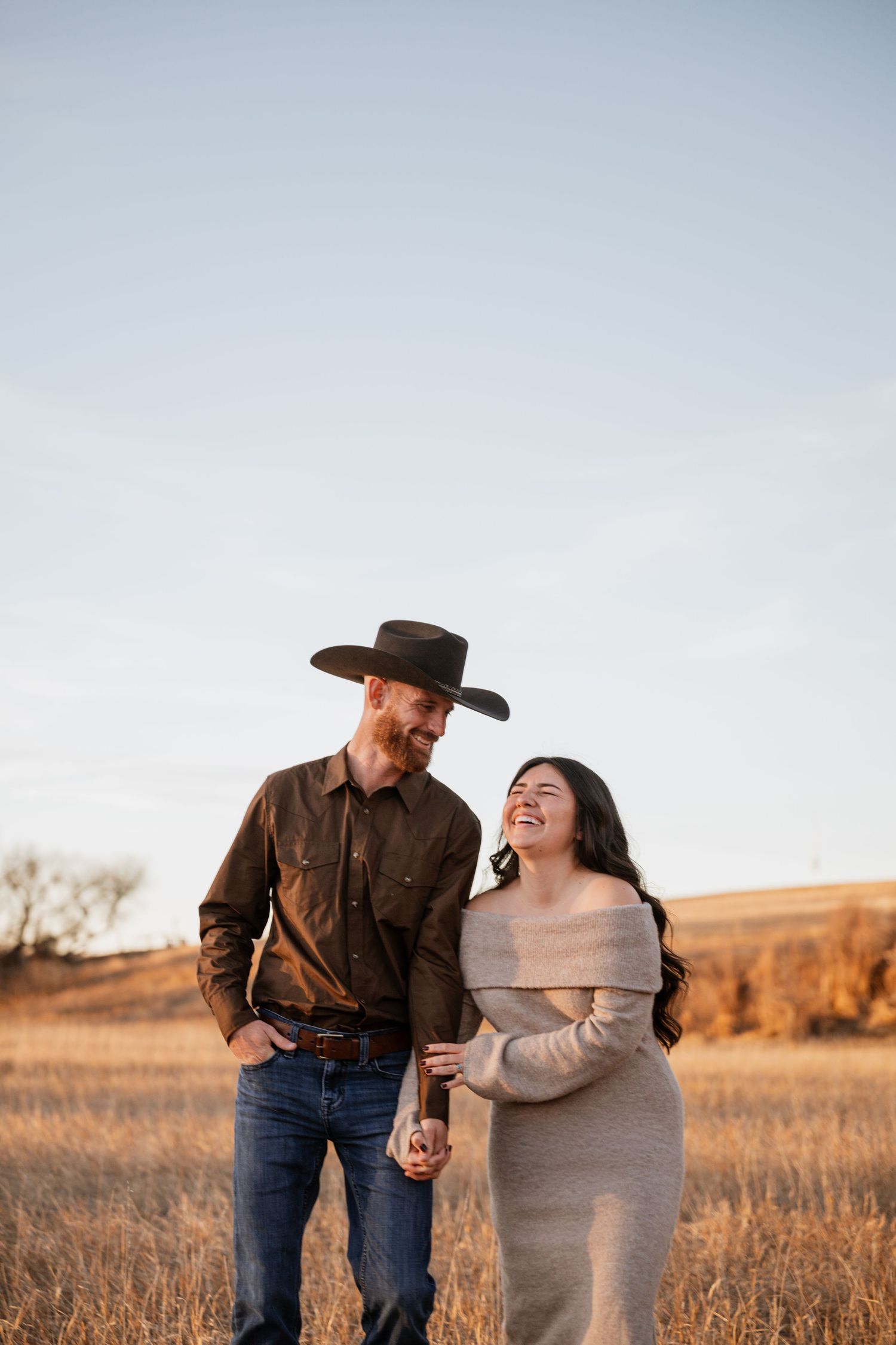 A couple shares a joyful moment together in a golden field during sunset, wearing western-style attire.