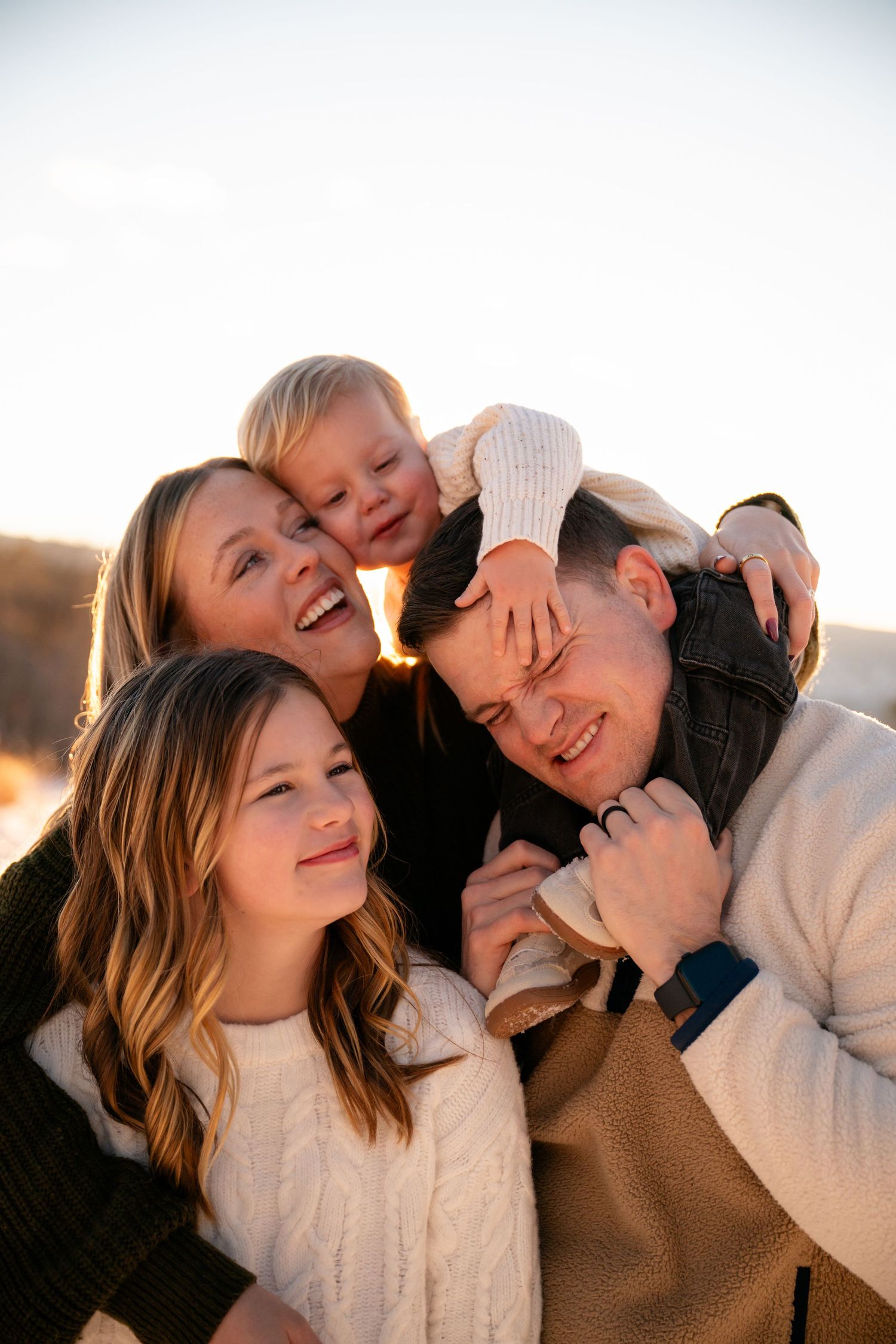 A group of people in cozy sweaters share joyful embraces and laughter during a sunset photoshoot outdoors.