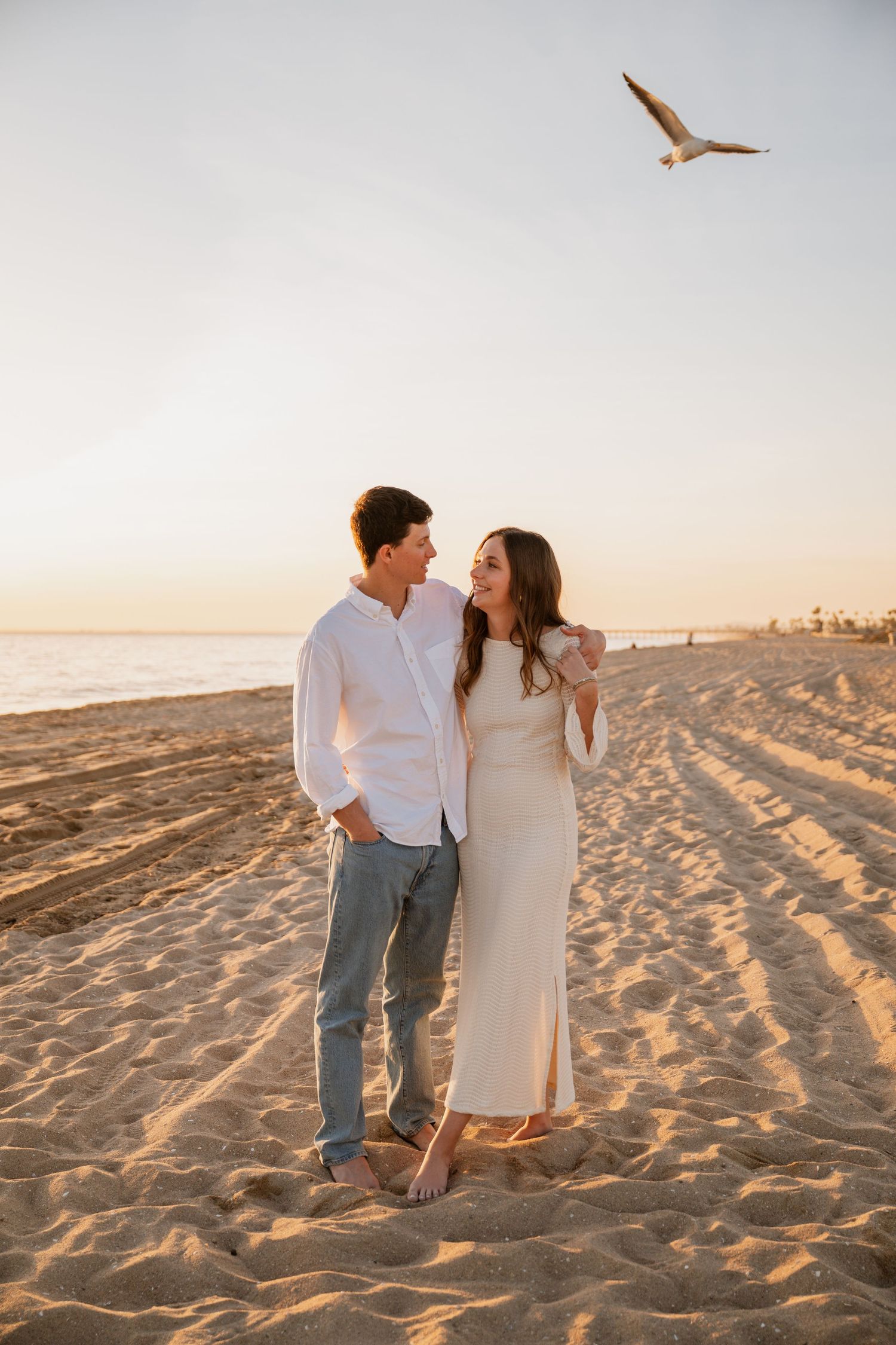 A couple in white outfits shares romantic moments on a sandy beach during golden hour sunset.