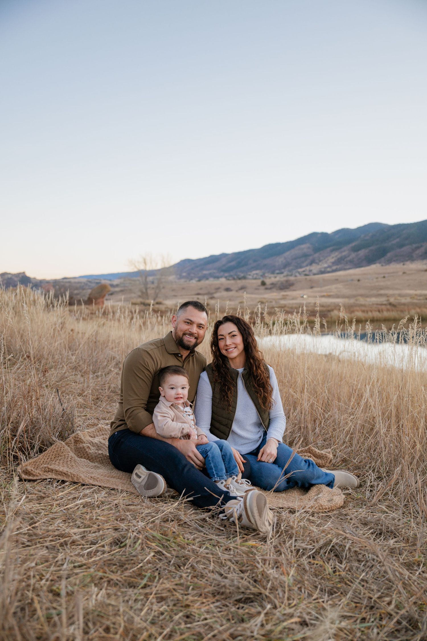 Family sits together in tall grass near a lake with mountains in the background during golden hour.